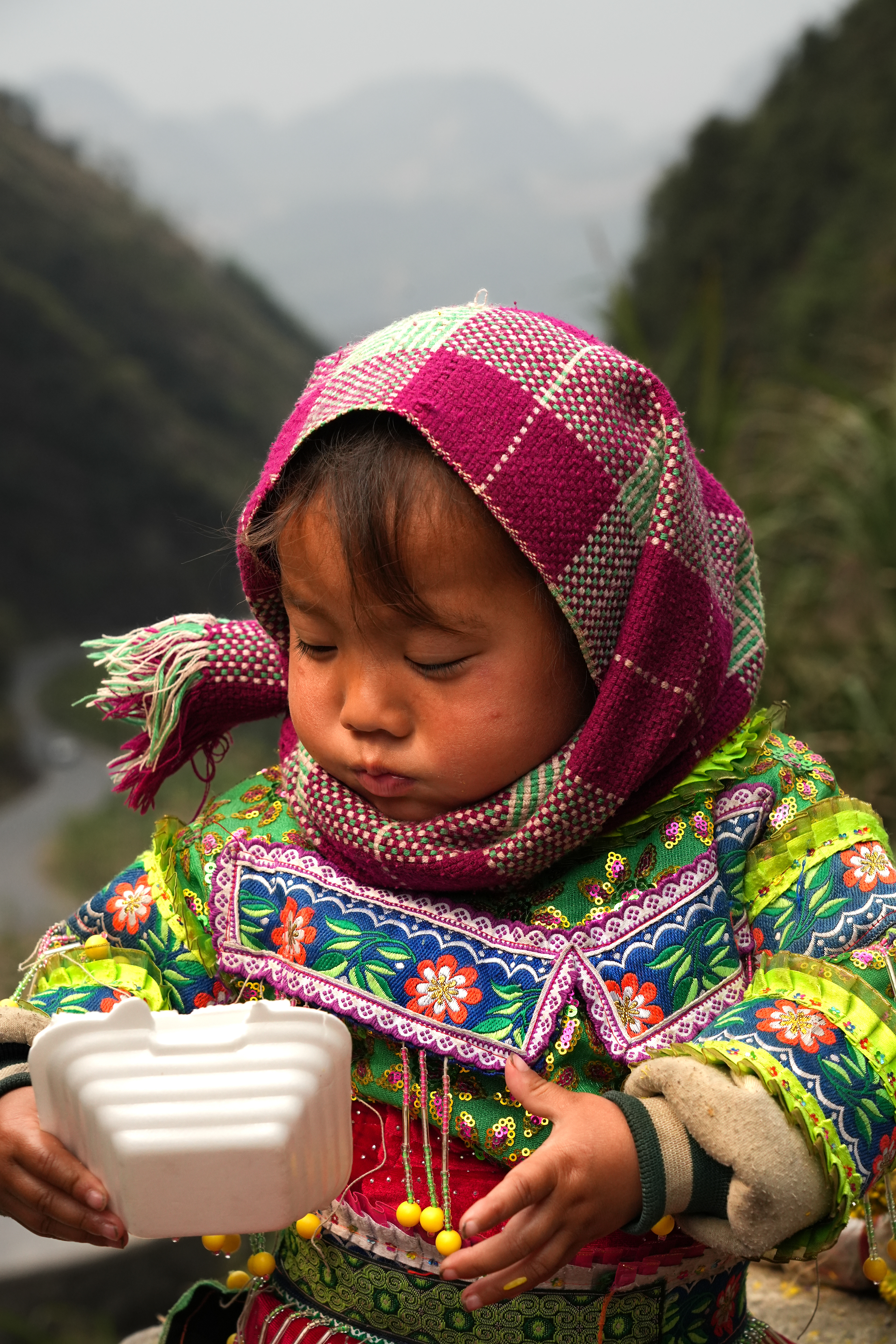 A Little Girl enjoying her lunch in front of a Mountain View in the Ha Giang province. What is she eating? Is it good? Her parents are around, trying to earn a living from tourists. Yet warnings are whispered among visitors: don’t buy flowers from these little girls, don’t let them braid your hair, sometimes they’re kept out of school because tourists are willing to pay for their labour. Do they make enough? Are they keeping her from school to earn more? Or is she simply too young to go to school? Do they come here every day? She is so small. How old is she? What do they call her? What is her name?