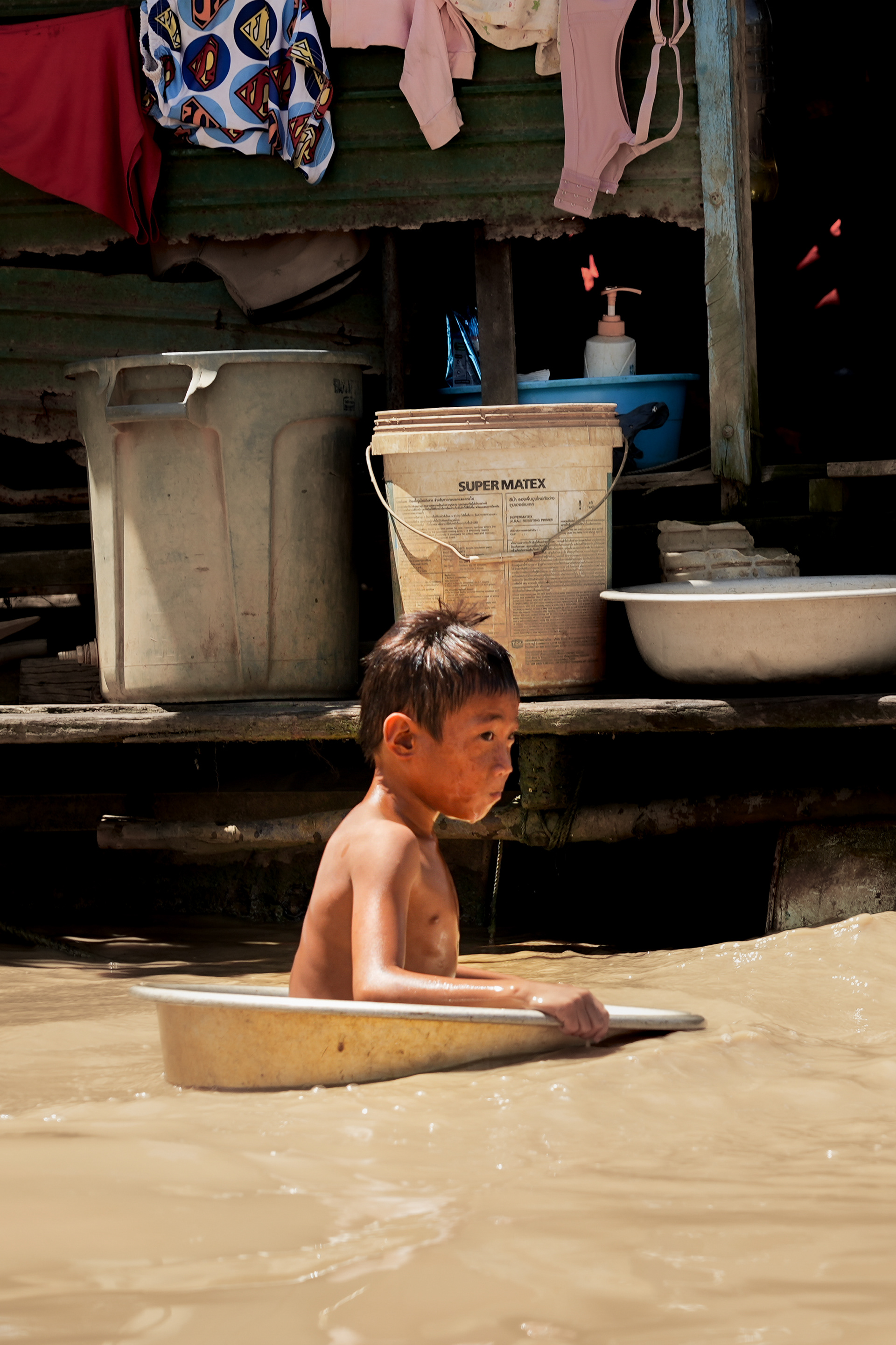 A little boy bathing in a soup dish in a fisher's village in Cambodia. His playing pool is where his parents fish their dinner. Is he playing in the soup dish or cooling down? Is he aware that his playing pool is the village’s main source of revenue? Does he know that the river is supposed to be blue? That soil turns the river brown? That dirt is meant to stay on the ground? Has he always been doing this? How old is he now? What is his name?