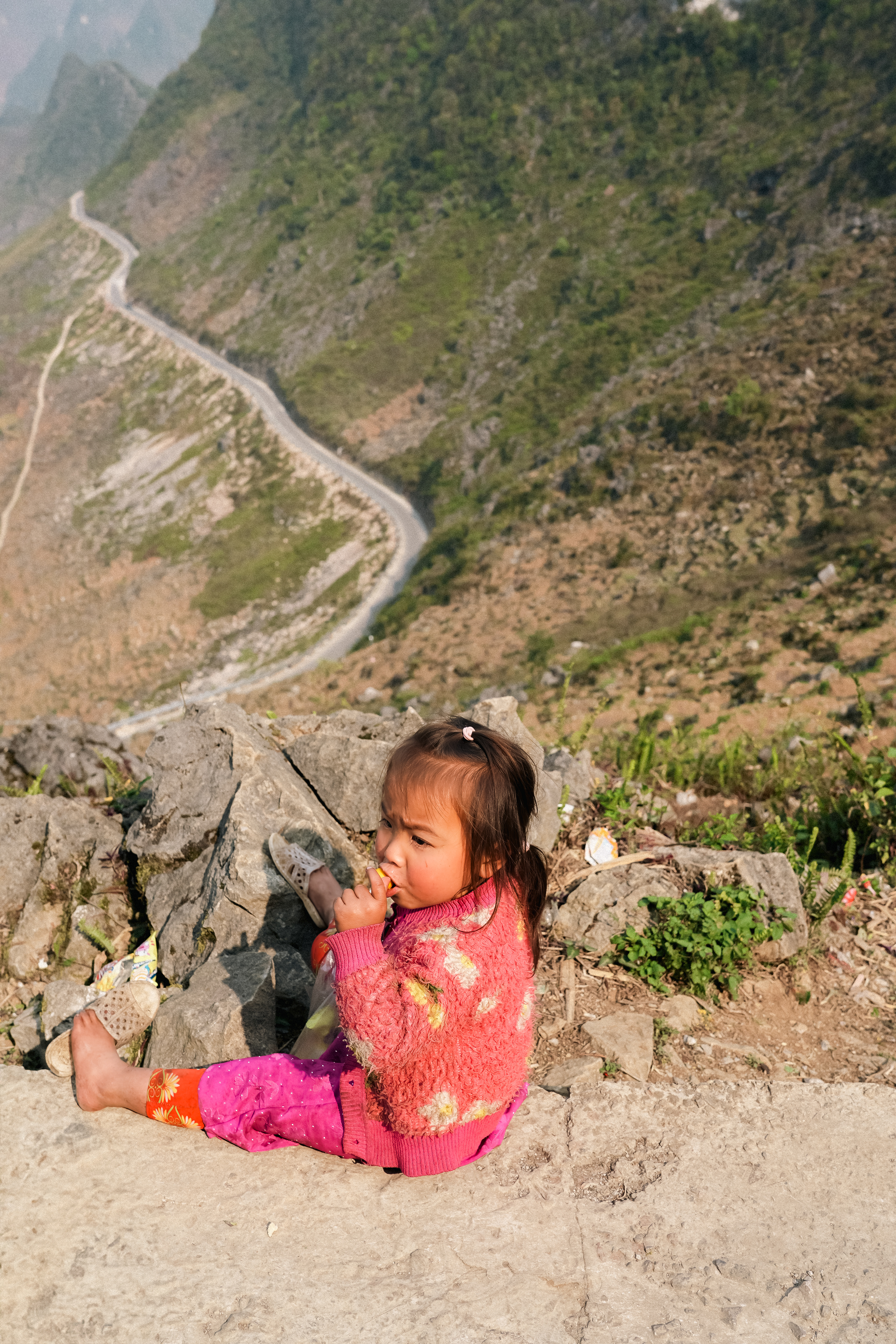 Vietnam. This astonishing destination. It is very famous to us, GenZ, backpackers, kids of the world. Among its top activities is the Ha Giang Loop, a stunning four-day journey through the mountains in northern Vietnam. Some people go for the thrill, some for the views, some for both. I went to explore the culture, to capture it. I knew the views would be incredible but I knew the culture would be more than that. With my camera in hand, that I was lucky to get on a deal, I found myself wondering how we reconcile exploring lives shaped by hardships with the comforts that make it possible. This Little Girl sitting on the edge of a mountain on the Ma Pi Leng Pass, in the Ha Giang province. She was sitting here, eating her lollipop while a trail of gen z kids were going back on their motorbikes, that they had paid a small fortune to be on. The edge of the mountain is rather scary. Is she safe sitting like this? Is this a real danger or only a danger to me, unaware of what life in the mountains is like? Does she even live in the mountains or only come here during the day? Where is the nearest village she could live in? Where are her parents? Are they watching over this young Little Girl? How old is she? What is her name?