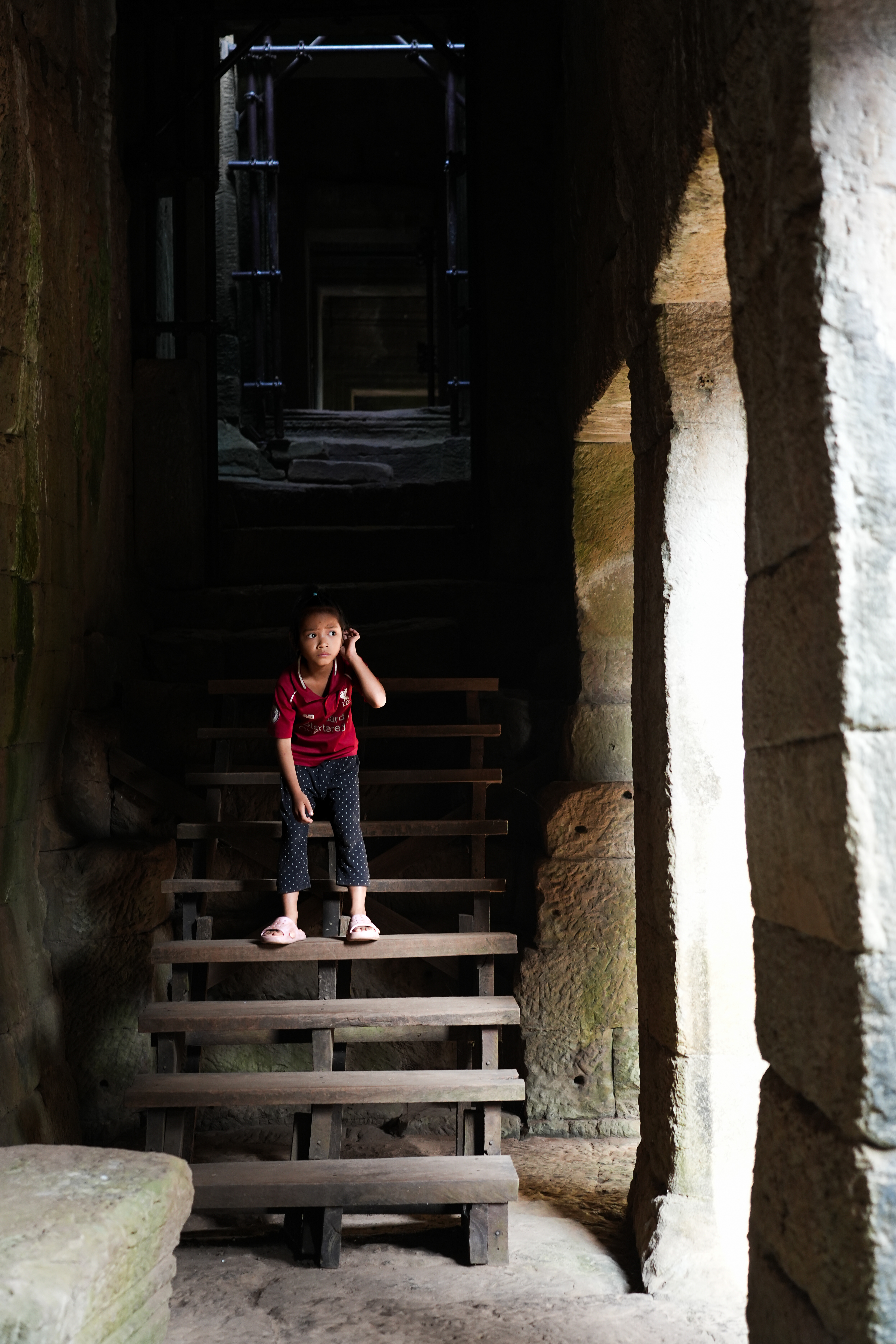 A Little Girl stands in Bayon Temple, Cambodia, her gaze unsettled, searching. Kids drifted in and out of sight, running through the arches, the shadows, and the light. Across the street, the religious ceremony was still taking place. Was it following Visak Bochea, an important Buddhist holiday that had taken place the day before? I assumed that these kids were playfully waiting for their parents. But she wasn’t running. She stayed still, looking lost, as if she was waiting. For what, or for whom, I cannot know. What is she waiting for? Who is she waiting for, her parents? Are they attending the ceremony across the street or working as tour guides? Is she waiting for them or waiting for someone else? Is she even waiting at all? Is she alone? Lost? Does she have siblings? Are they nearby? Should we let someone know she is here? How can we contact them? How would we call them? What are their names? What is her name?