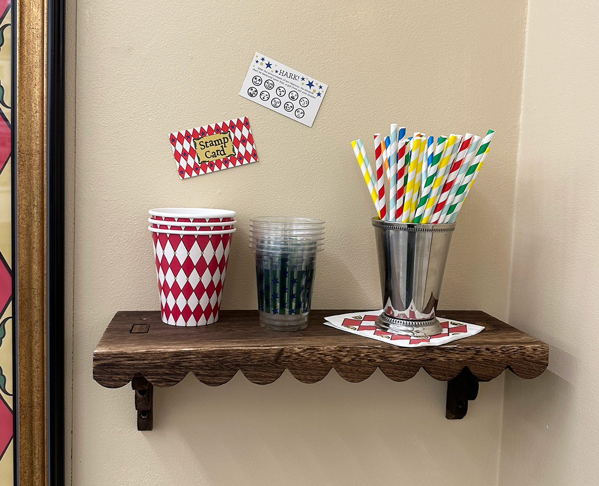 Left shelf used to display extra cups, the front and back of the stamp card, a silver cup that holds paper straws on top of a napkin.
