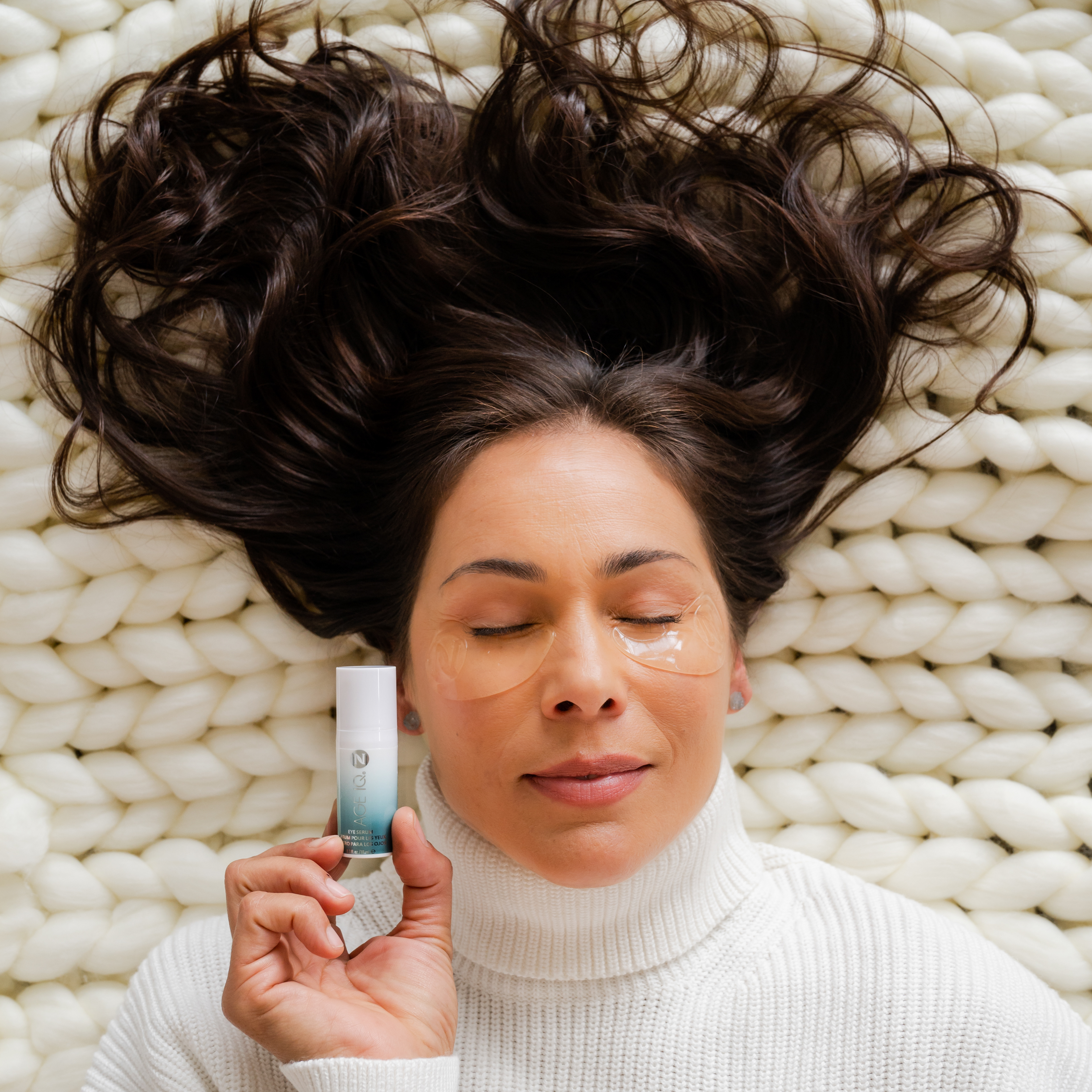 Woman laying on a woven blanket with eyes closed. She is wearing underage patches and holding Neora Age IQ next to her face.