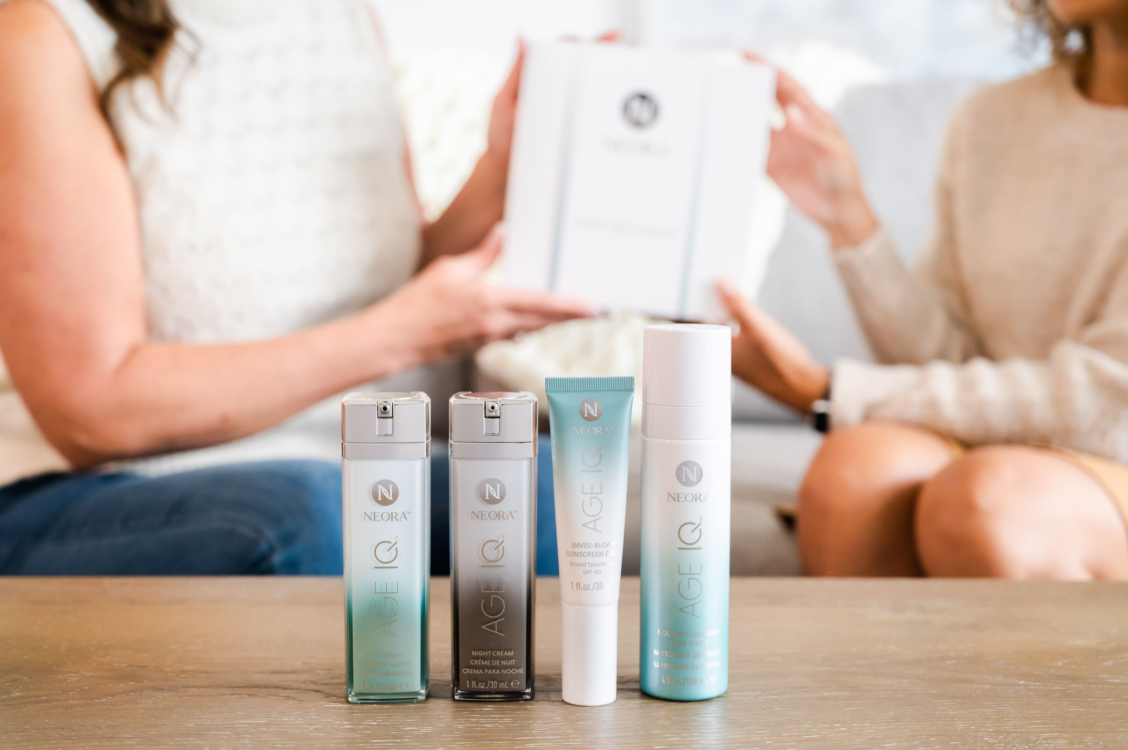 Close up shot of Neora Age IQ product line with ladies exchanging gifts in the background.