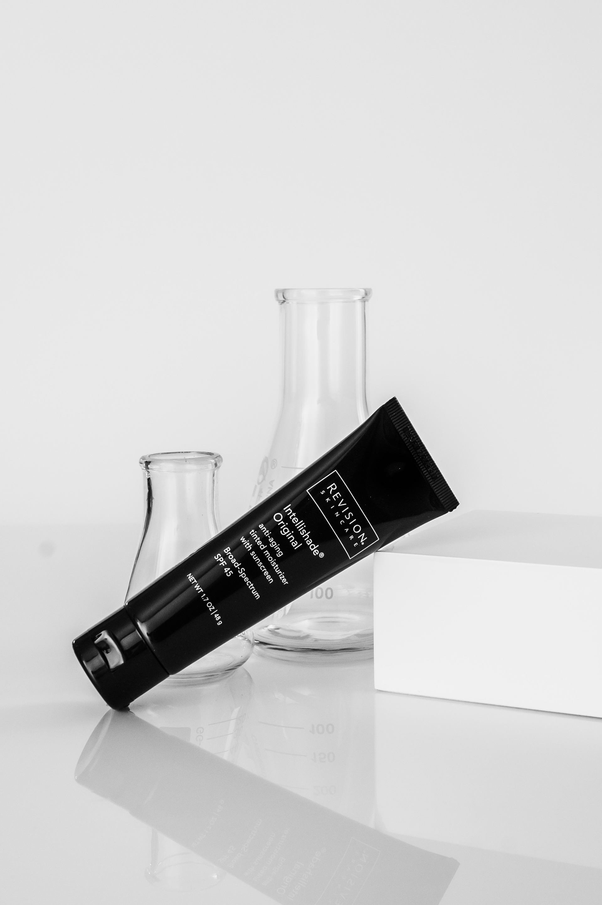 Black and white image of Revision Skincare Intellishade SPF styled leaning against a white block with chemistry flasks in the background