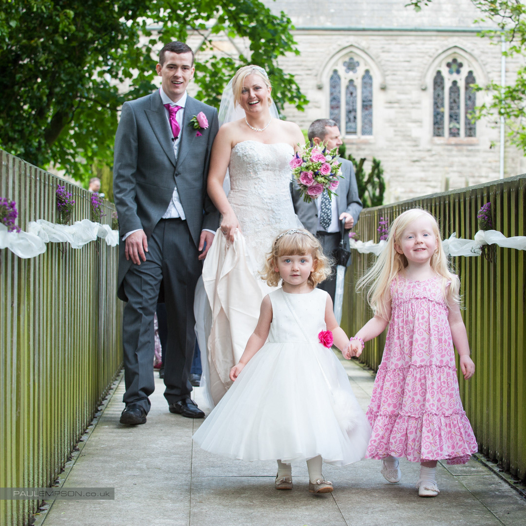 Flower girls hand in hand lead a laughing bride & groom over a bridge at North Yorkshire Wedding