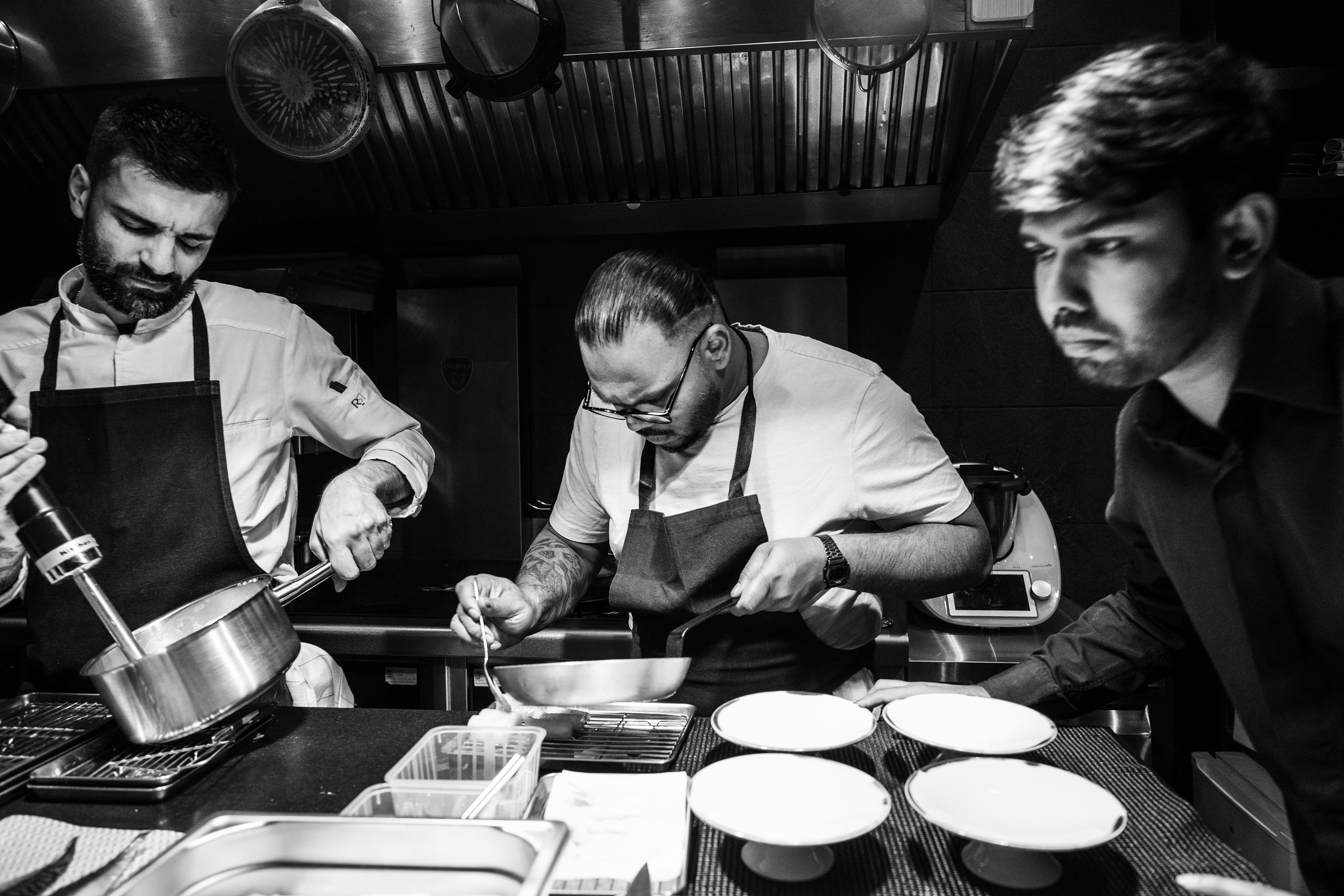 On the left, kitchen assistant Vincenzo is blending a mixture, whilst in the centre chef Manogeran Shasitharan is piping a mousse onto artichoke hearts, and on the right a waiter is preparing to take the dishes out to the dining room, on 26 March 2026, at Mantra restaurant, Paris (9th arrondissement), France.