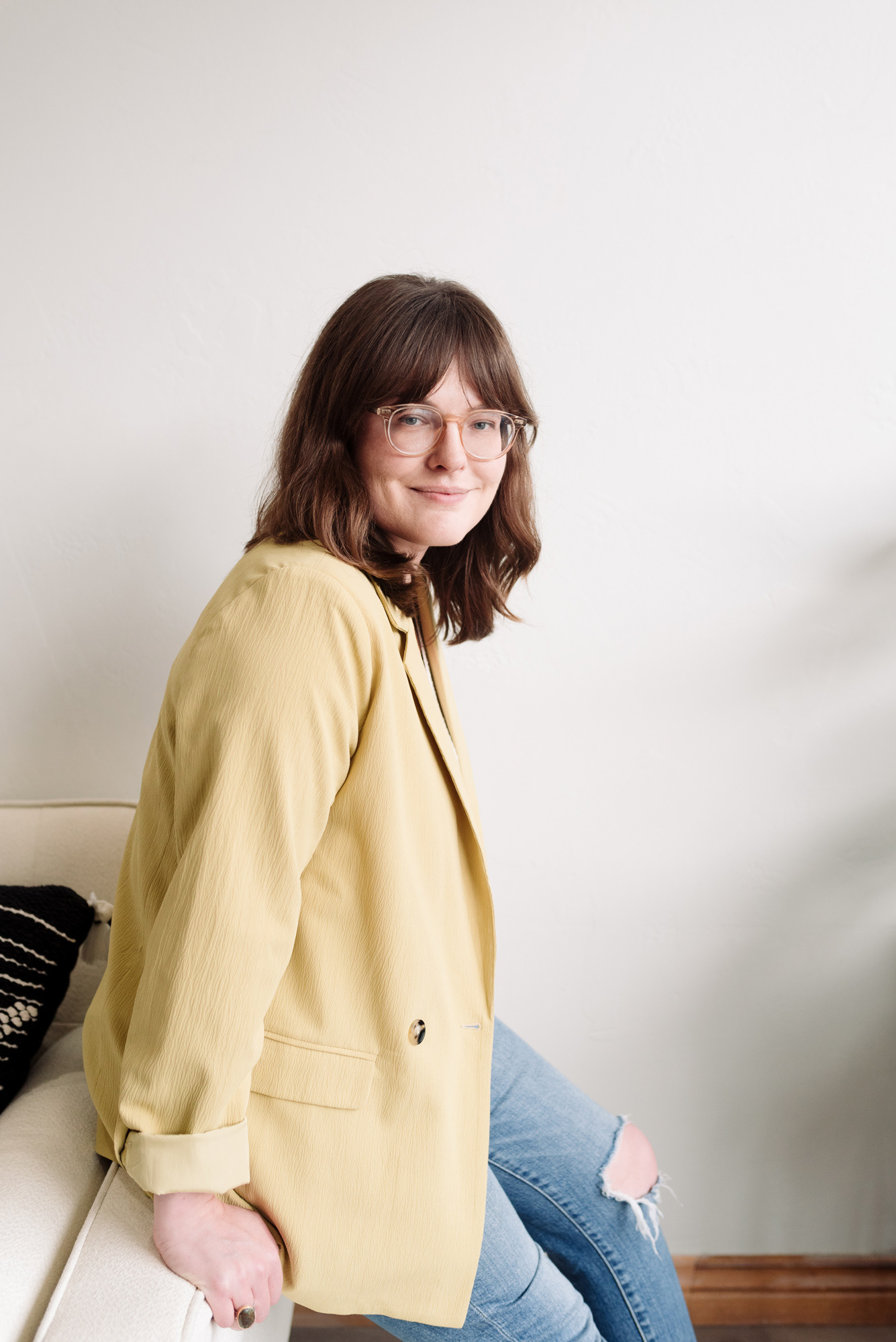 Portrait of a woman sitting on the arm of a sofa and leaning forward in jeans and a yellow jacket.
