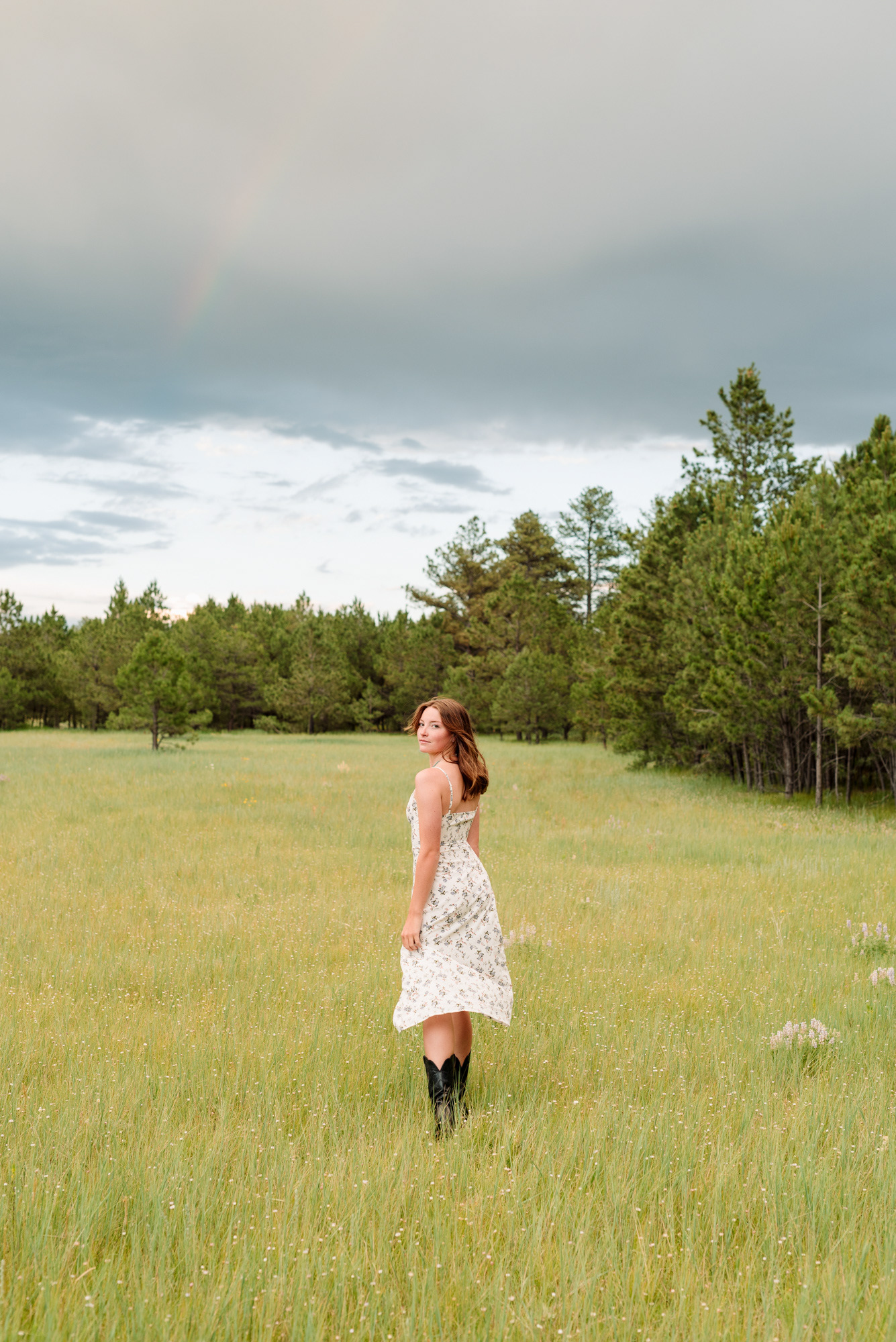 Senior portrait of a girl in a field in Colorado walking away from the camera with a rainbow in the sky..