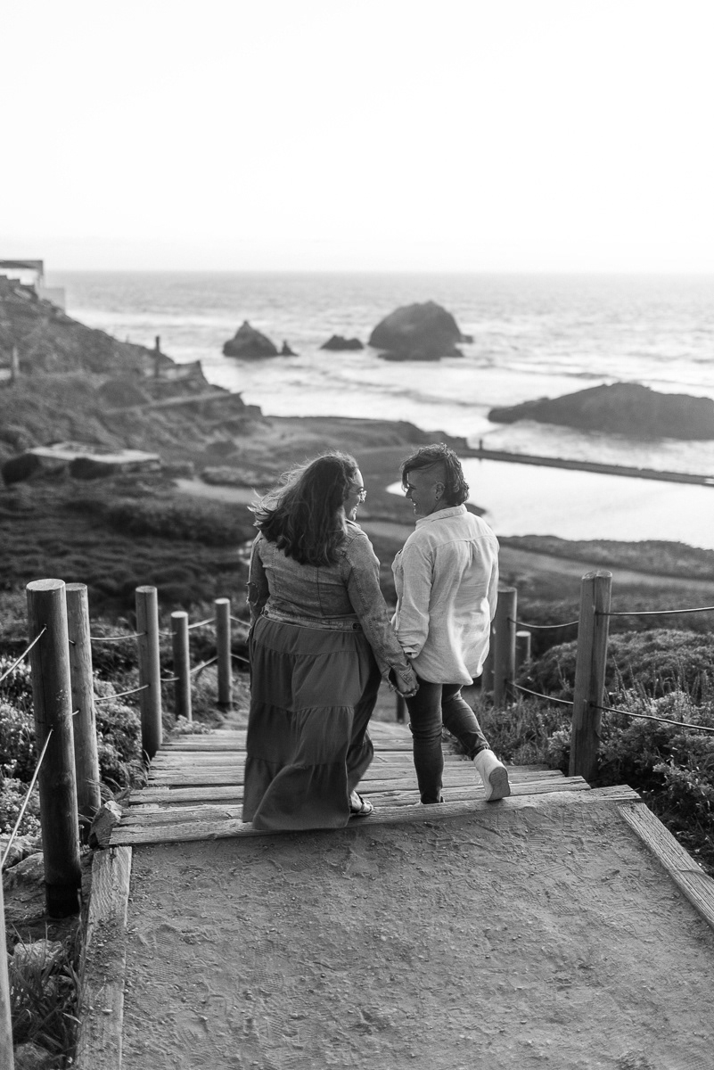 Two women walking down stairs at the beach front looking at each other and holding hands.