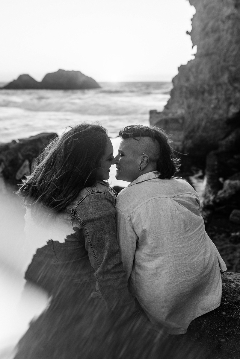 Two women sitting on rocks at the beach leaning in for a kiss.