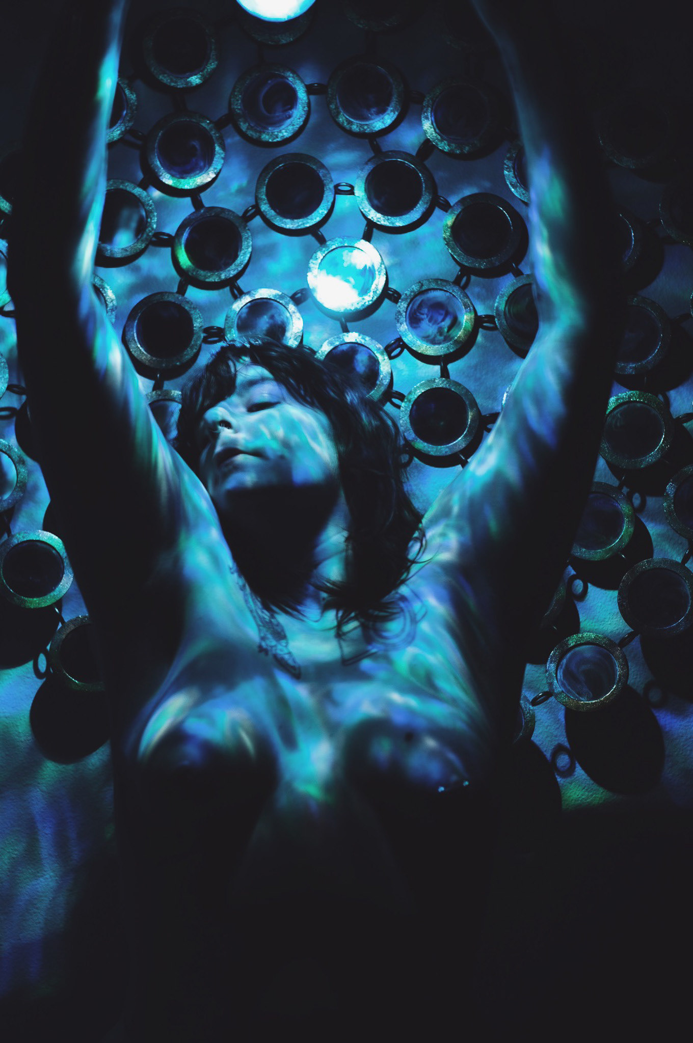 Image description: Sami was sitting on the bench topless and her arms were up in the air. There was a galaxy projector on everything. It was in a blue tone.  There was a mirror behind her.