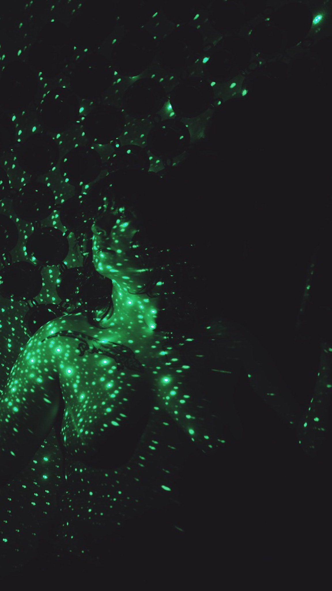 Image description: Sami was topless sitting on the bench against the wall in her room. The green stars projector was on the wall including Sami. Sami was looking the left.