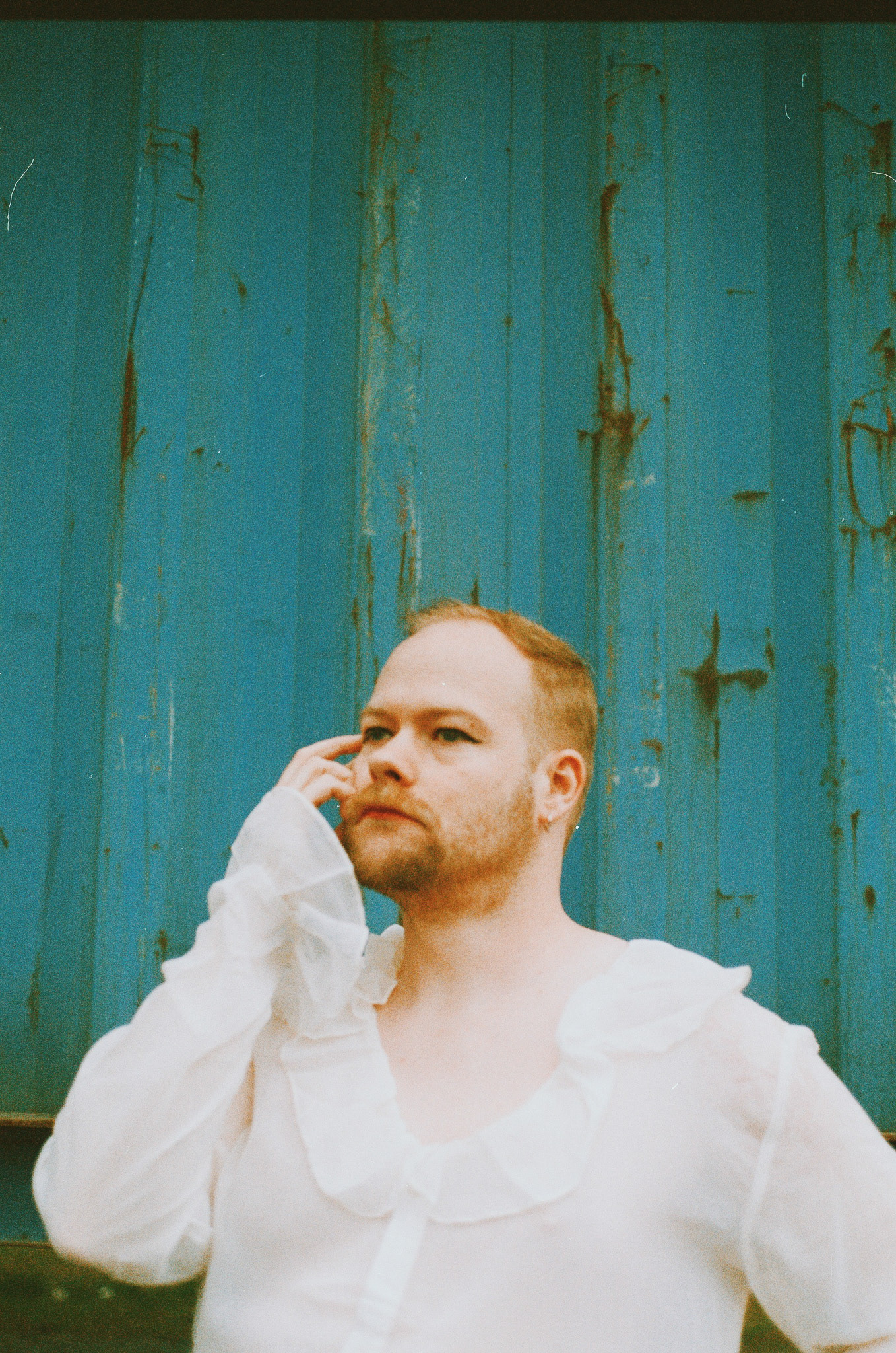 Image description: the color film portrait of a person with a red beard and red hair standing in front of a teal container, wearing a fluffy white top.