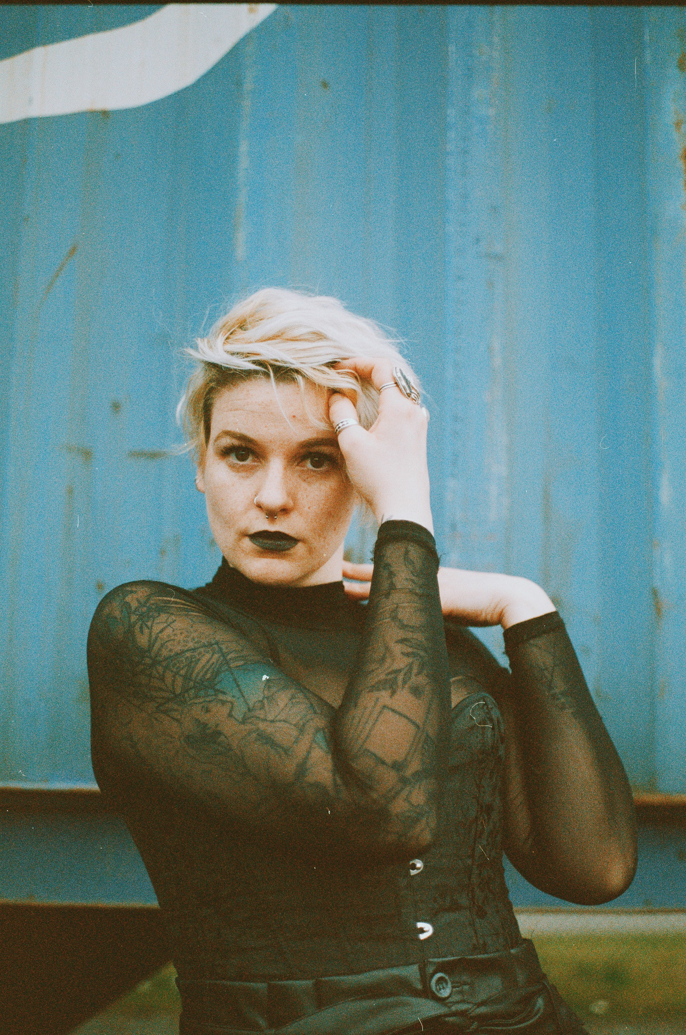 Image description: a woman wore black lips, a black lace top, and blonde short hair.a woman standing with her hands on his head. She is in front of a container wall.