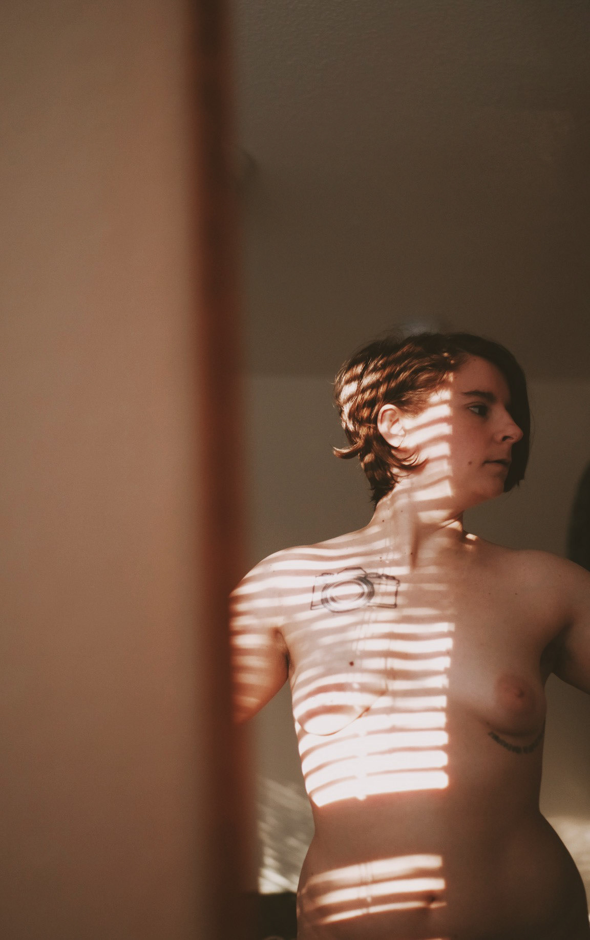 Image description: The mirror reflection was Sami naked in her bedroom. There was lighting through the curtain on her face, chest, and shoulder.