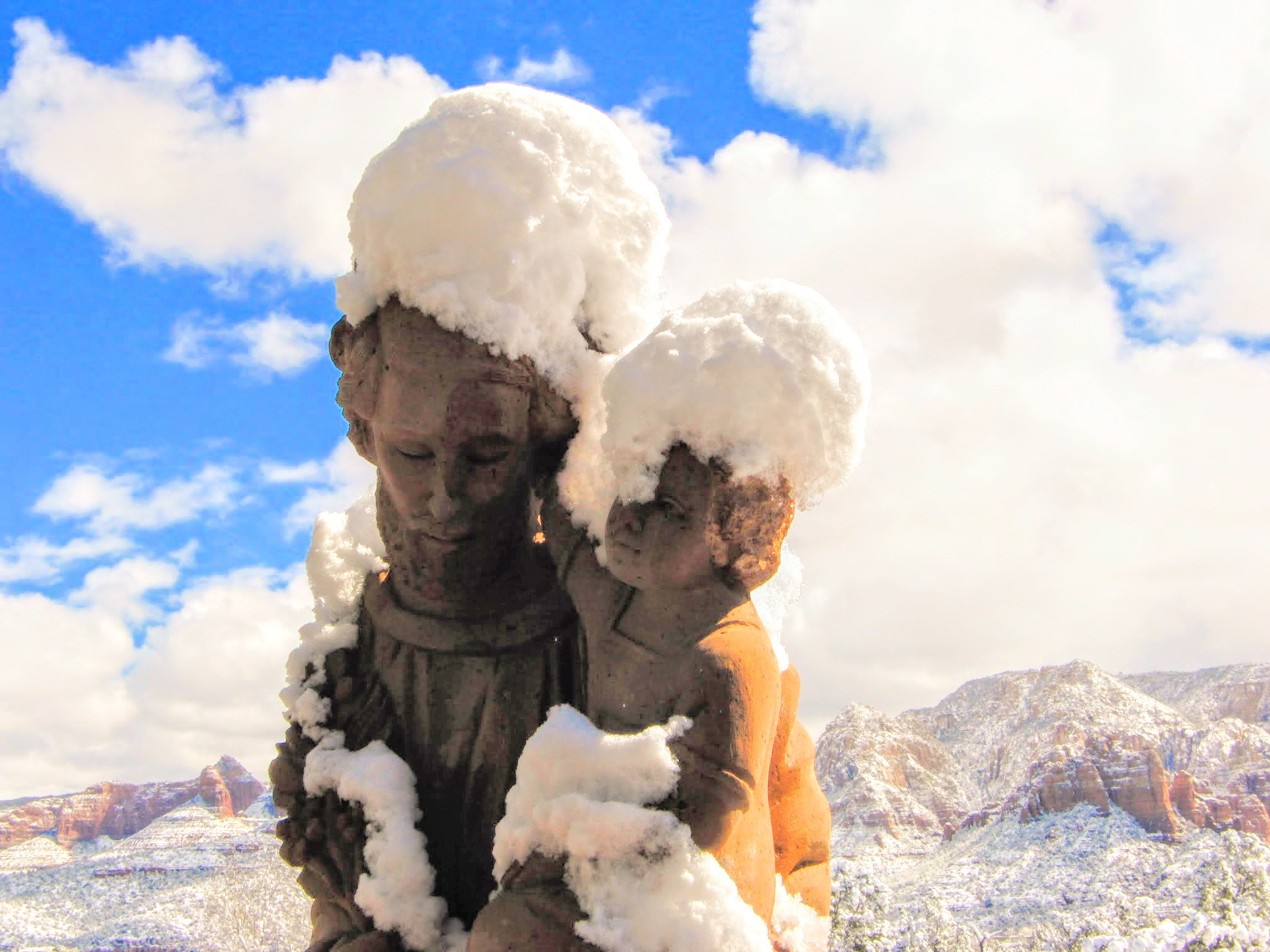 The image captures a serene winter moment in the picturesque setting of Sedona, Arizona. It depicts a sculpture of a father carrying his young son, both adorned with whimsical hats of snow, a playful touch from nature. The surrounding landscape is a tapestry of contrasting colors: the vibrant red rocks, distinctive of Sedona's famous geological features, peek through a delicate dusting of white snow, symbolizing the rare and gentle touch of winter upon this desert scenery.  The sculpture conveys a sense of protection and support, a visual metaphor for the father's role as a carrier and guardian through life's challenges. It's a poignant representation of the times when one needs support, the figure of 'The Father' here becomes a universal symbol of parental love and sacrifice. As if the father in the sculpture is not just carrying the child through the snowy landscape, but also through the trials and tribulations of life.  This quiet yet powerful scene evokes a feeling of nostalgia and warmth amidst the cold, a reminder of the enduring strength of familial bonds and the comforting presence of a parent's unwavering support.