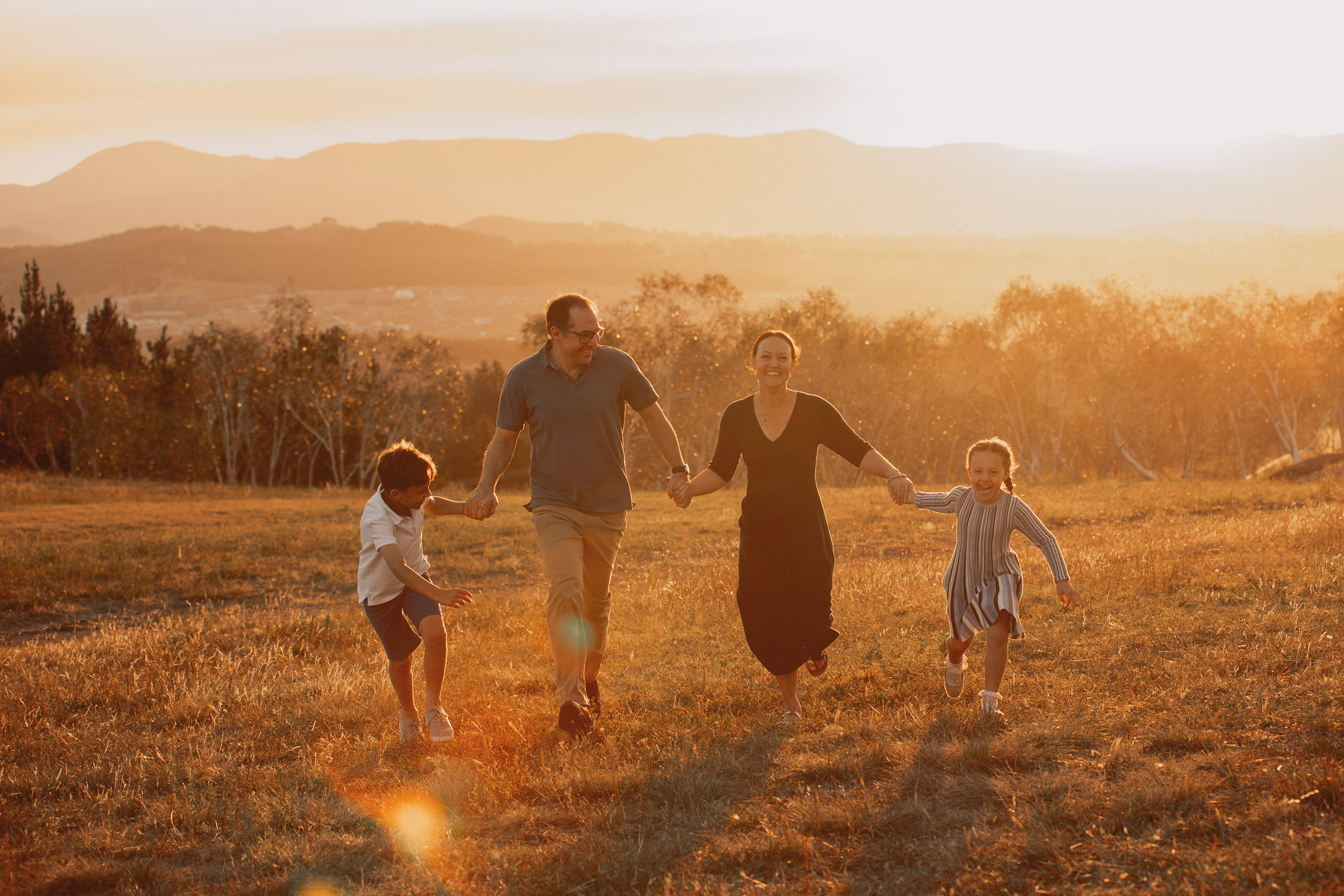 A happy family of four running hand in hand through an open field at golden hour, surrounded by warm sunset light and rolling mountain views. The golden sun creates beautiful lens flare and soft backlighting, capturing a joyful outdoor family photography session filled with connection, laughter, and natural moments.