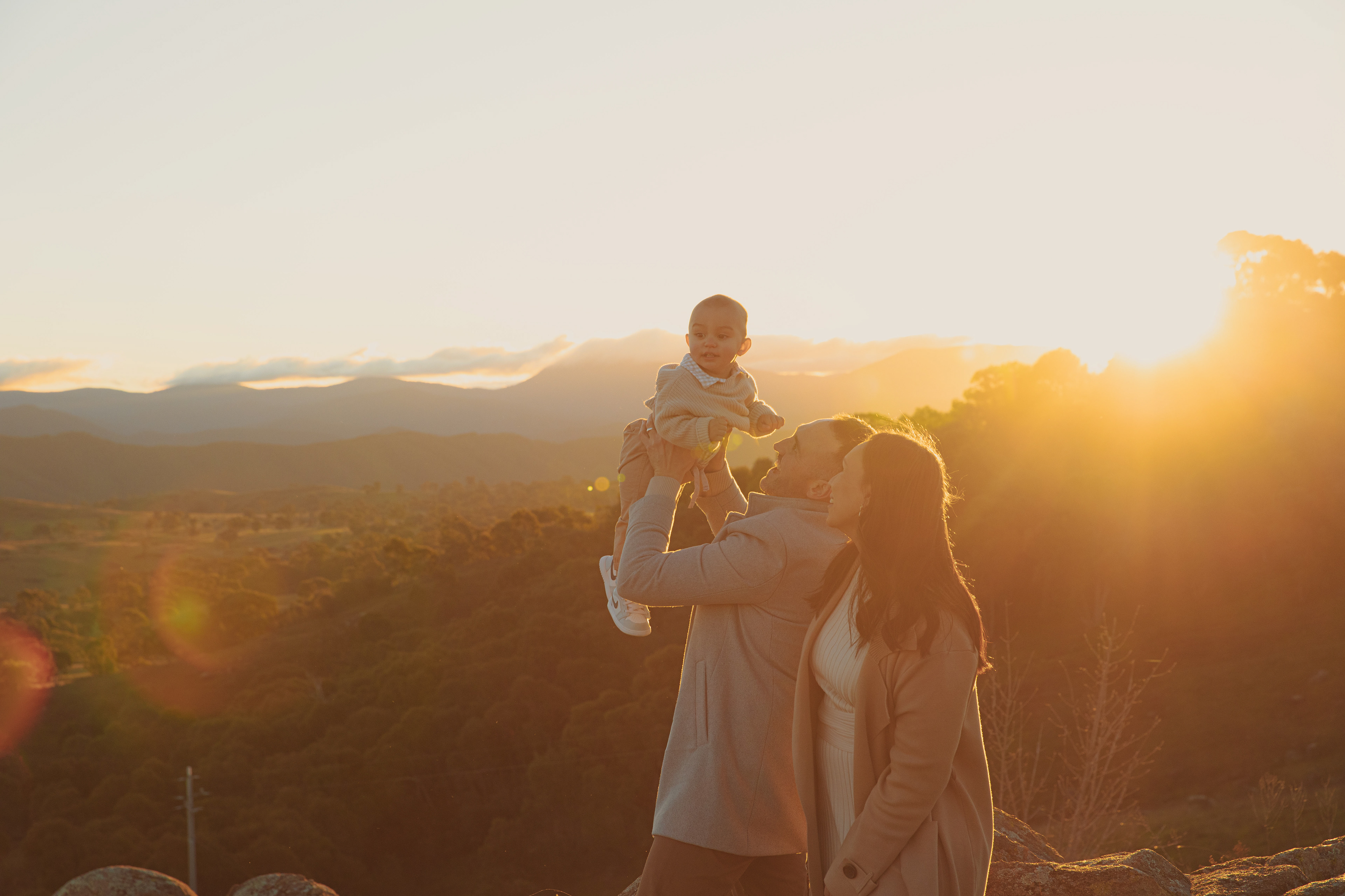 Family enjoying a golden hour sunset in the mountains, with parents lifting their baby into the warm evening light. Captured during sunset photography, the image features glowing sun flares, soft golden tones, and a breathtaking mountain landscape, creating a timeless family portrait filled with warmth, connection, and natural beauty.