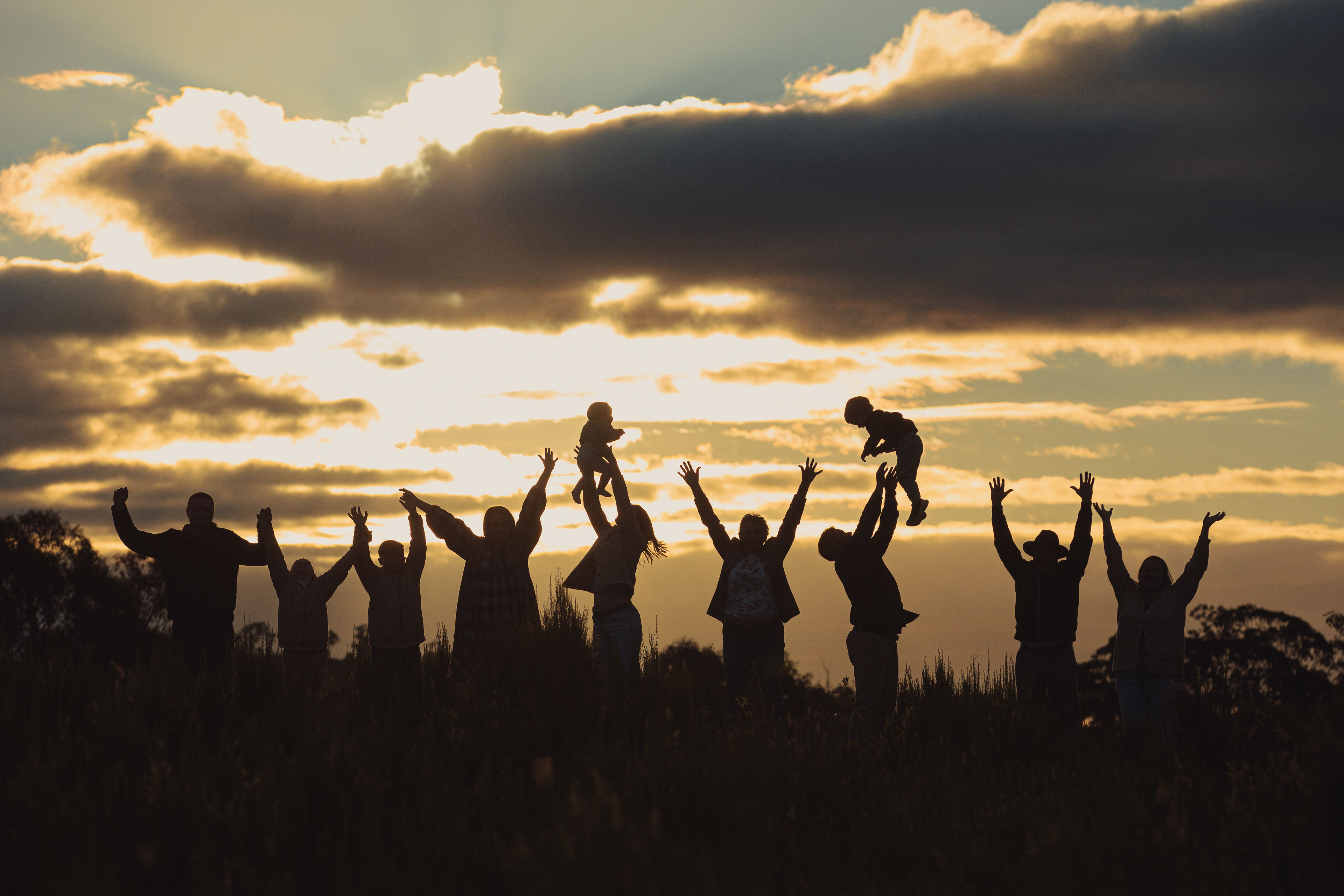 Extended family silhouetted against a golden sunset, celebrating together with raised hands and children lifted into the air.