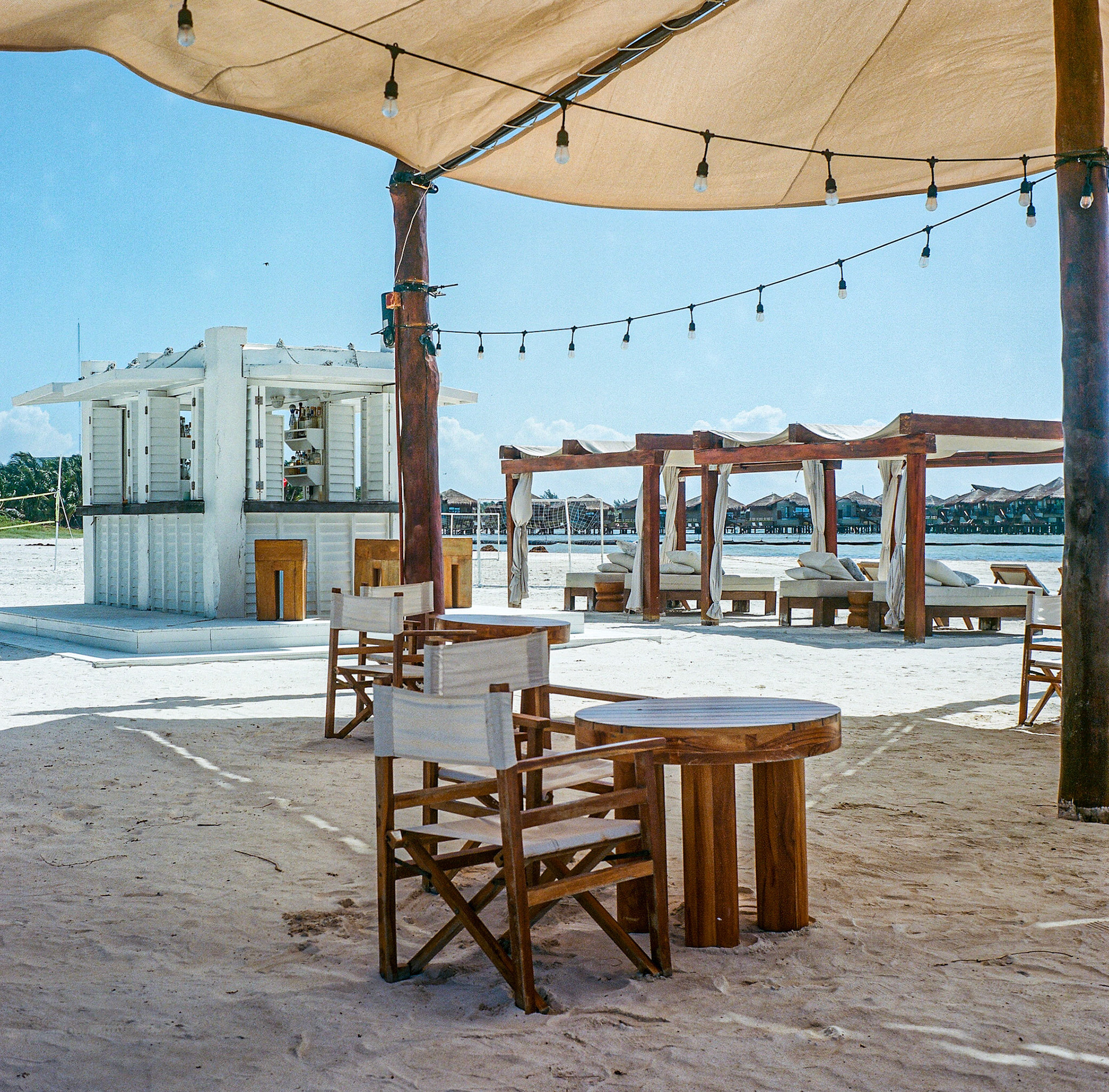 Beachfront dining area with wooden tables, umbrellas, and string lights beside the ocean. Explore the full series at dyoon.space.
