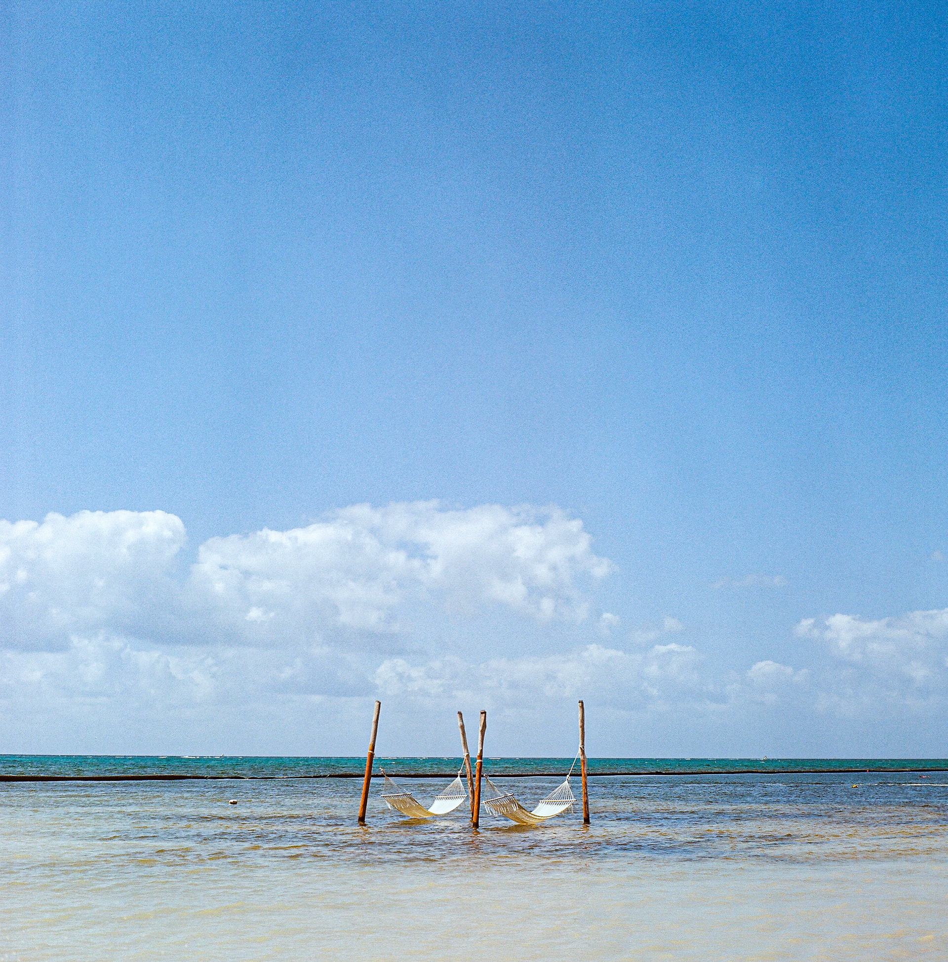 Hammocks suspended above shallow turquoise water beneath expansive blue skies and soft clouds. Explore the full series at dyoon.space.