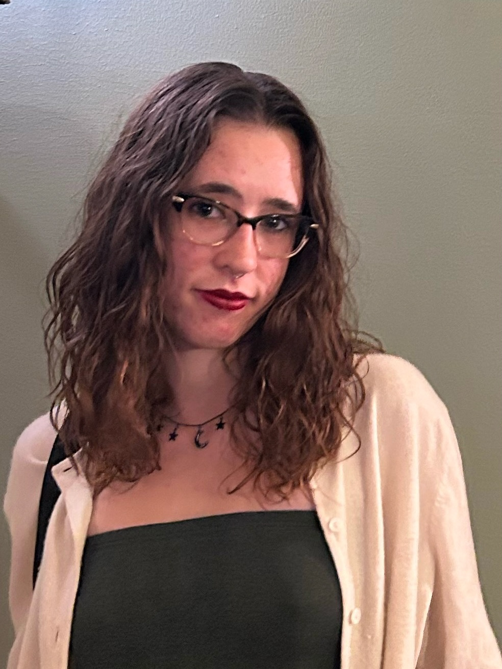 Headshot of Denali Norton, smiling at the camera with her hair down. Wearing a white sweater, a dark green top, a black moon necklace, and glasses in front of a light green backdrop.