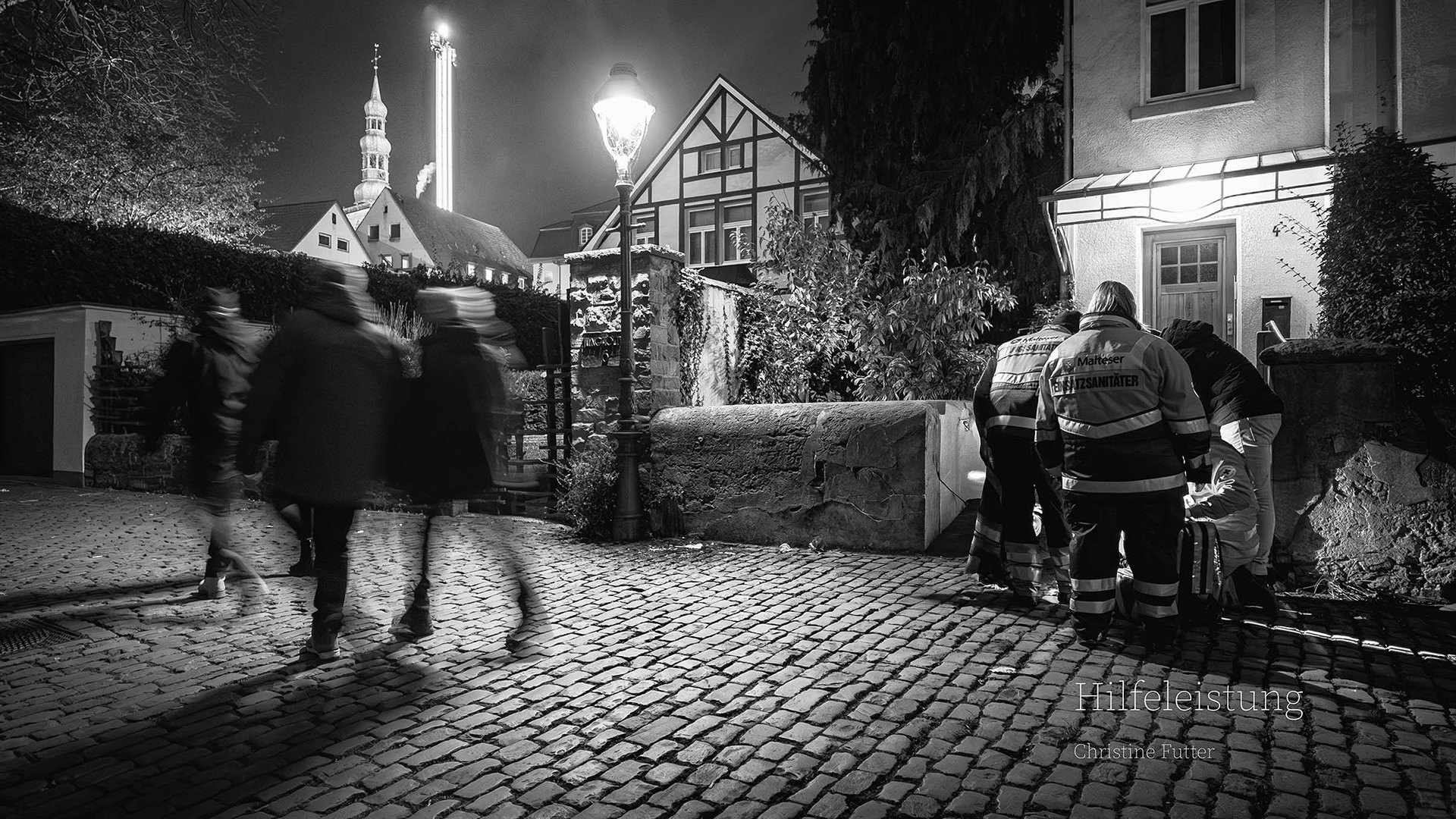 Parallelwelten - Ein Sanitätstrupp kümmert sich in einer Gasse um einen Menschen, Kirmesbesucher eilen vorbei. Über den Dächern sieht man den Turm eines Fahrgeschäftes und einen der Soester Kirchtürme. Zwei sehr gegensätzliche Bauwerke über zwei sehr gegensätzlichen Menschengruppen.