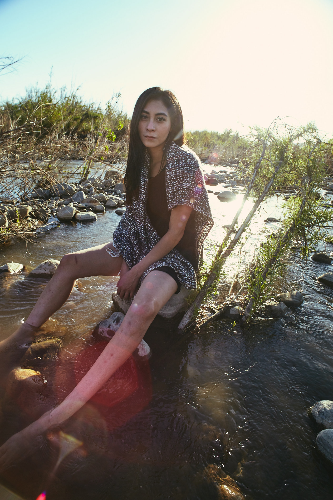 Beautiful woman sits on rock in creek bed.  Water streams.  Photo by Modern Human