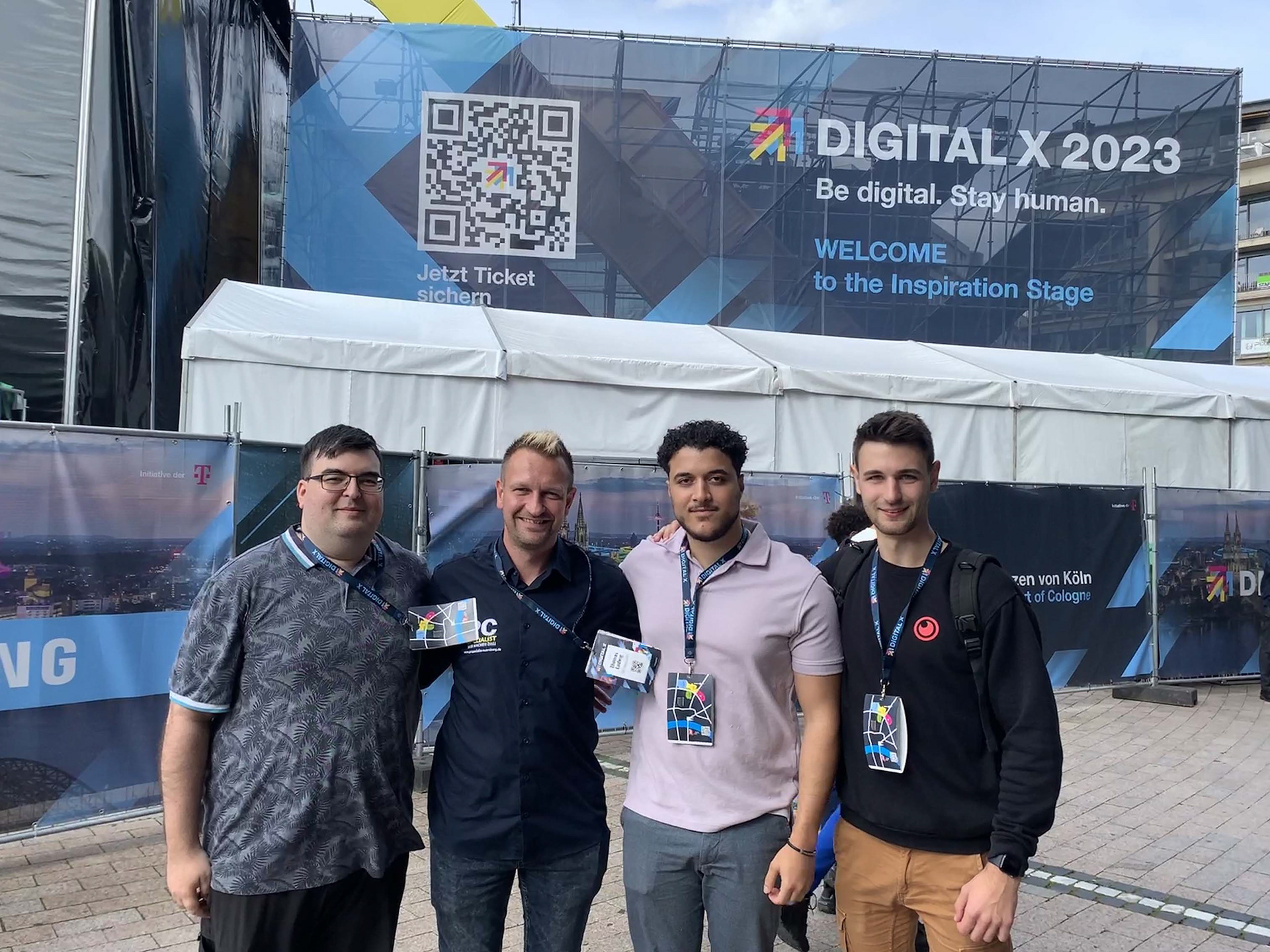 Das Bild zeigt vier Männer vor einem Banner der "DIGITAL X 2023" mit dem Slogan "Be digital. Stay human." und einem QR-Code für Tickets. Sie tragen Veranstaltungspässe und stehen draußen vor einem Zelt, was auf eine Technologieveranstaltung in Köln hinweist.
