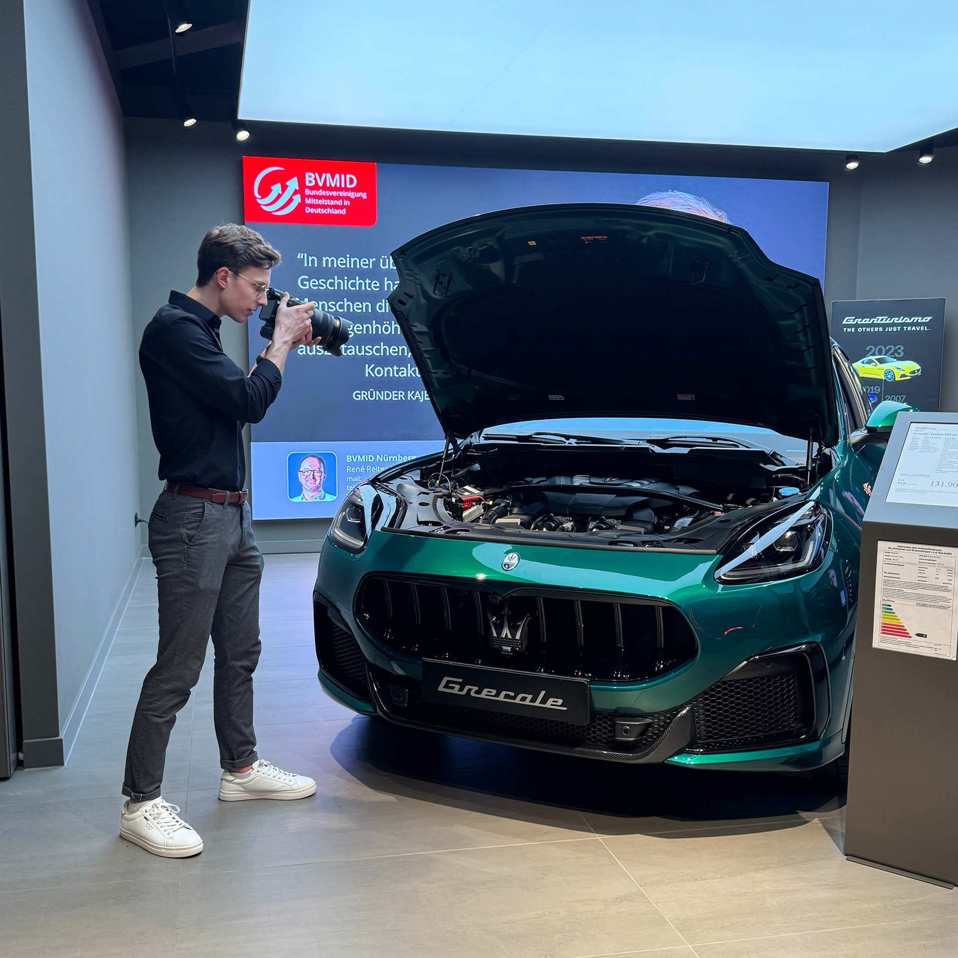 Ein junger Mann fotografiert ein grünes Auto mit geöffnetem Motorraum in einem modernen Ausstellungsraum. Im Hintergrund ist ein großer Bildschirm mit dem Logo und einem Zitat der BVMID zu sehen. Das Auto ist ein Maserati Grecale.