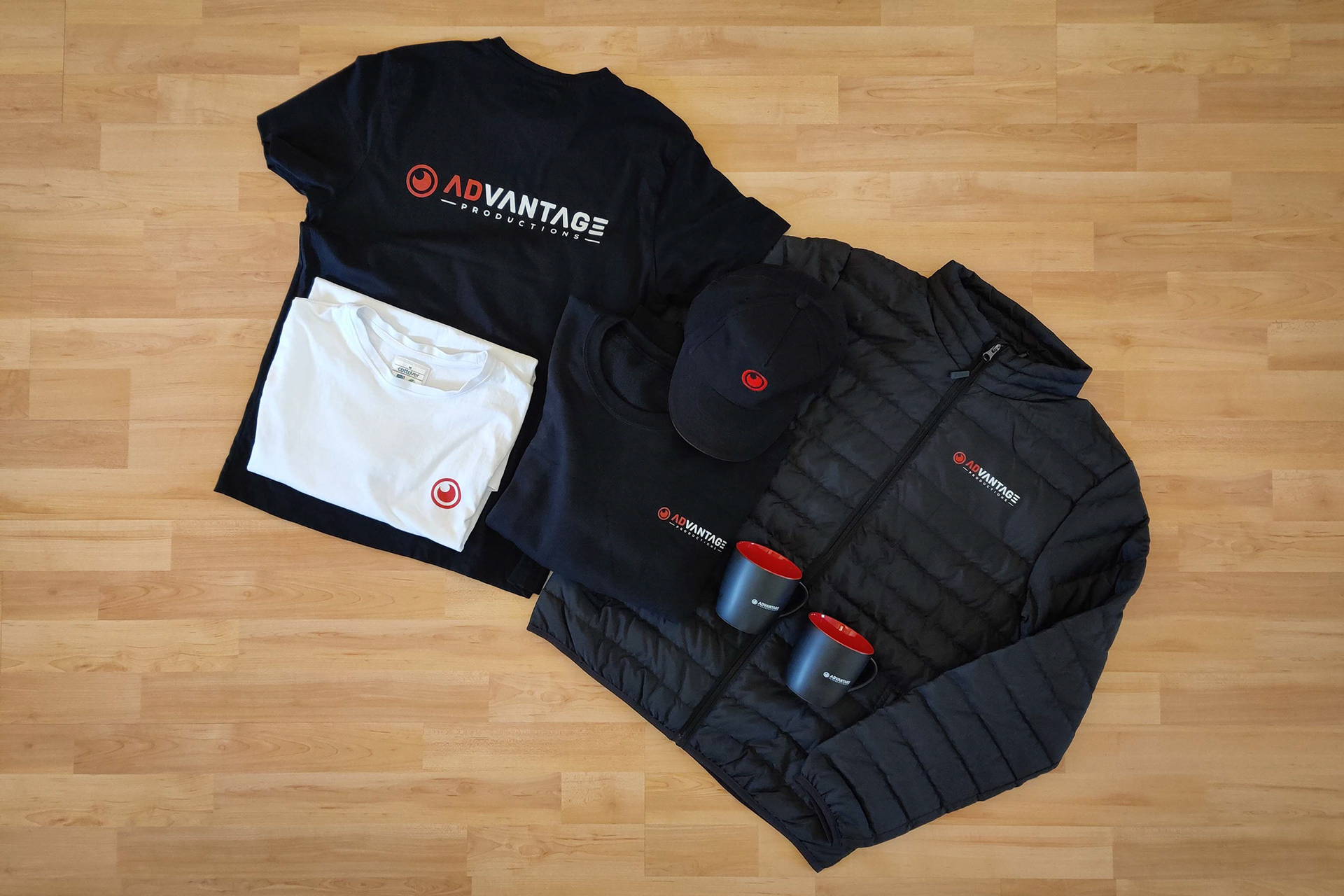 Schwarze und weiße ADvantage Productions Kleidung, einschließlich T-Shirts, Jacke, Mütze und zwei Tassen, auf einem Holzboden arrangiert.