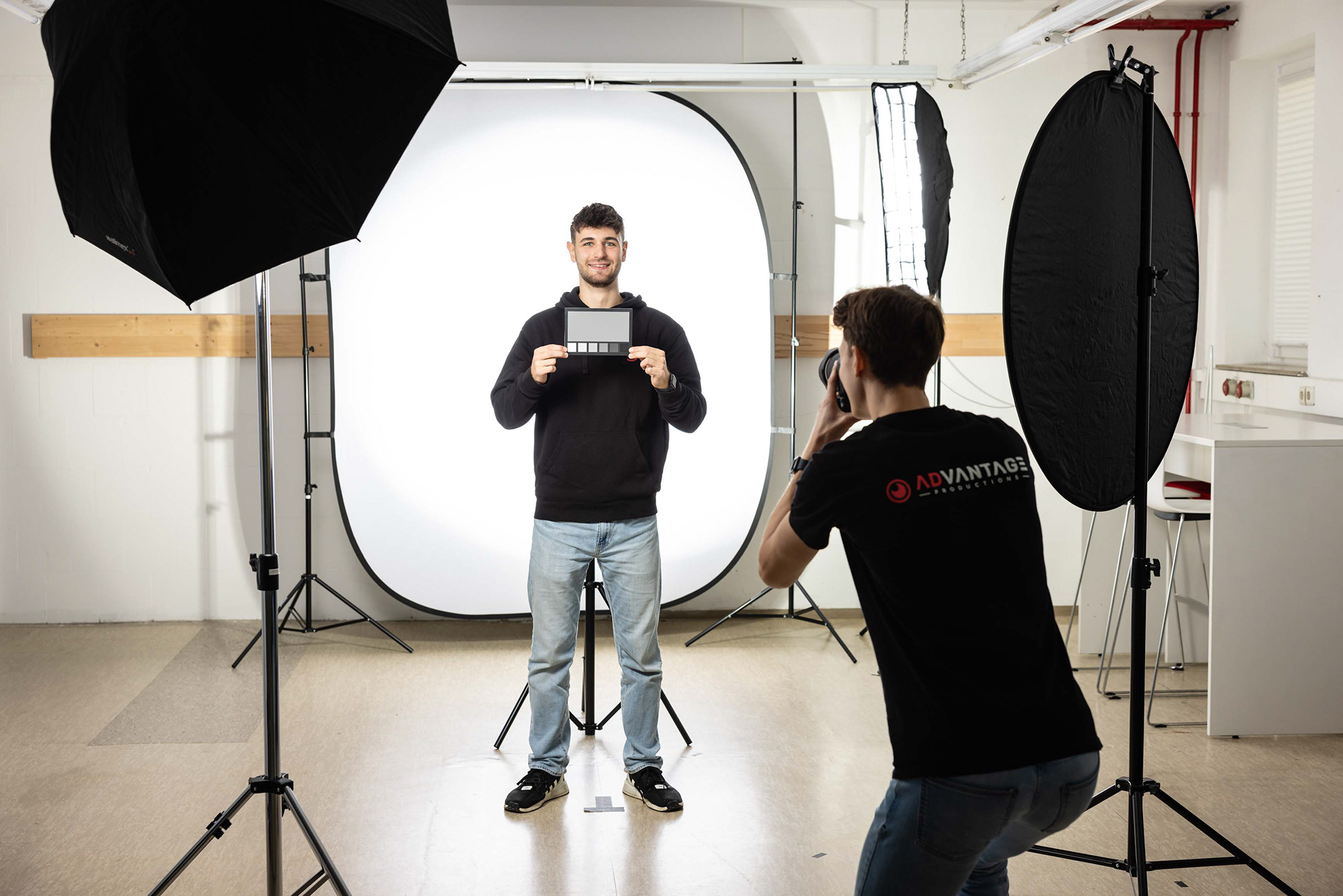 Das Bild zeigt eine Fotosession in einem Studio. Ein Fotograf von ADvantage Productions macht ein Foto von einer Person, die vor einem weißen Hintergrund steht und eine Graukarte hält. Um die Person herum sind mehrere professionelle Studiolichter und Reflektoren aufgebaut, um die Beleuchtung zu optimieren. Die Szene wirkt gut vorbereitet und professionell.