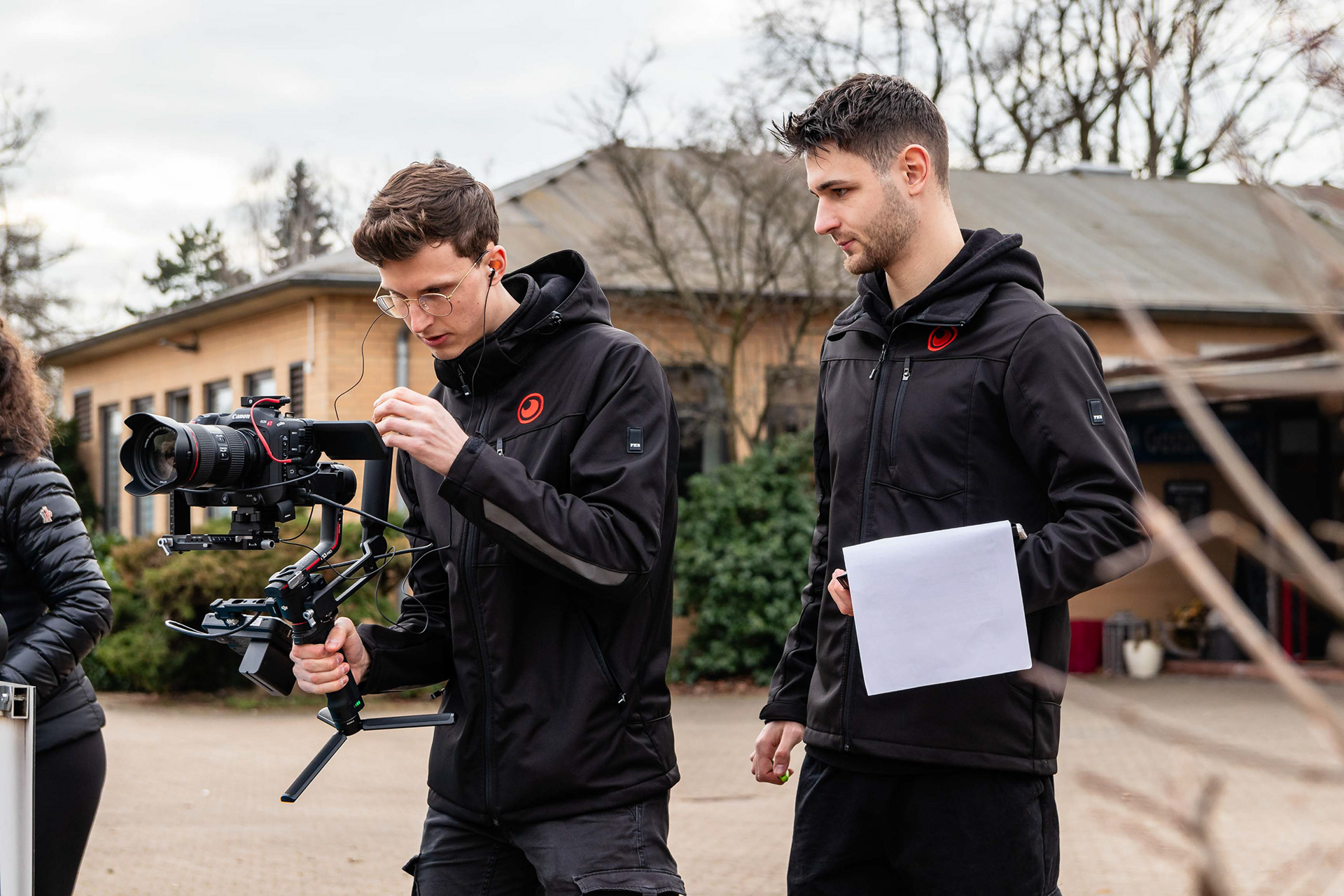 Zwei junge Männer in schwarzer Arbeitskleidung arbeiten draußen an einem Videodreh. Einer hält eine Kamera auf einem Stabilisator, während der andere ein Klemmbrett hält. Im Hintergrund sind Bäume und ein Gebäude zu sehen.