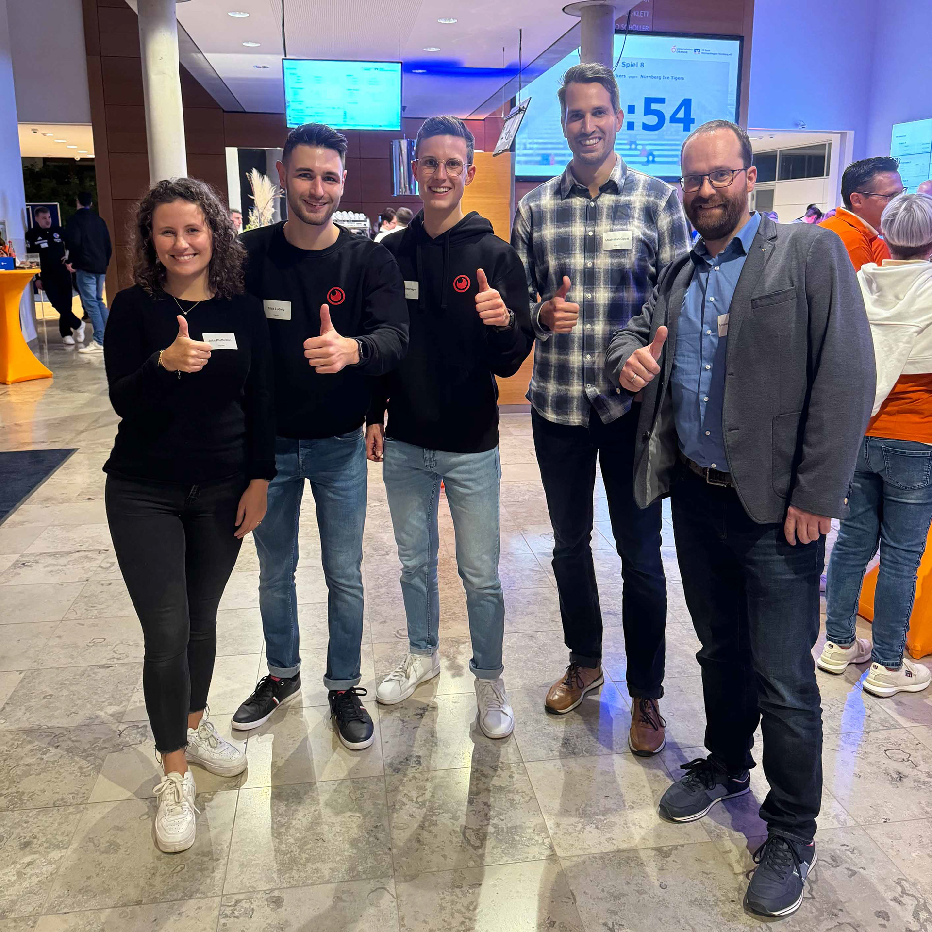 Das Bild zeigt fünf Personen, die in einer Innenumgebung Daumen hoch zeigen und Namensschilder tragen. Sie stehen vor einem digitalen Bildschirm, der Veranstaltungsinformationen anzeigt. Die Atmosphäre wirkt freundlich und entspannt.