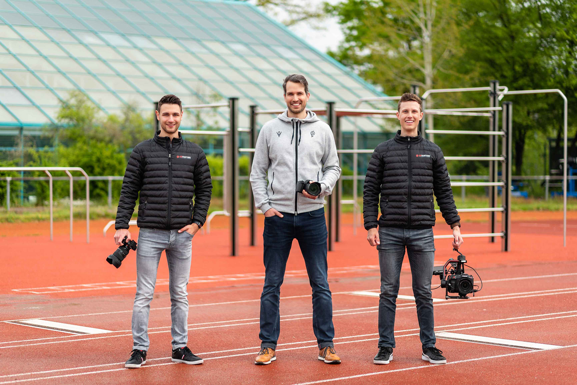 Drei Männer stehen auf einer Laufbahn im Freien. Zwei von ihnen tragen schwarze ADvantage Productions Jacken und halten Kameras, der Mann in der Mitte trägt eine graue Jacke und hält ebenfalls eine Kamera. Im Hintergrund sind eine Glasstruktur und grüne Bäume zu sehen.