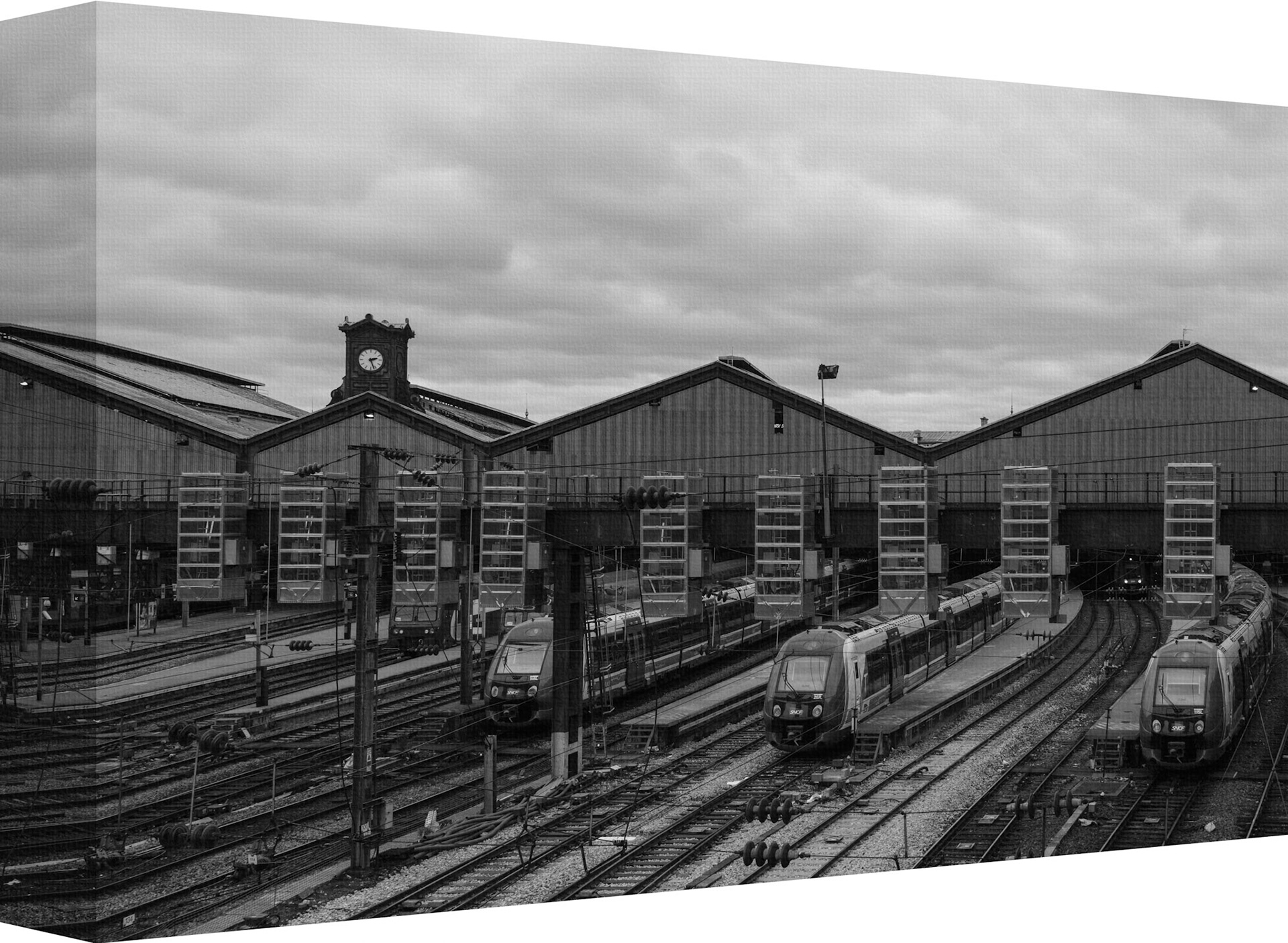 "Train Stop" is a captivating black and white photograph that offers a glimpse into the bustling world of railway transport. The image showcases multiple trains at rest, lined up on parallel tracks which lead the eye towards the grandeur of an industrial train station. The station itself boasts a classic architectural style with prominent roofing and a central clock tower that stands as a testament to timeless design amidst modern machinery. The overcast sky adds to the moody atmosphere of the scene, suggesting a moment of stillness in an otherwise dynamic environment. This photograph not only captures the essence of rail travel but also evokes a sense of nostalgia through its monochromatic palette, making it an intriguing piece for both art enthusiasts and train aficionados.