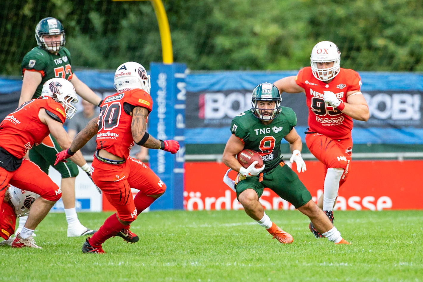 9 Aaron Bruning (grün) gegen 9Roni Salonen, 30 Jamaal White und 93 Julian Völker (von links rot) im Spiel der Kiel Baltic Hurricanes (grün) gegen die Braunschweig New Yorker Lions (rot) am 03.08.19 im Kilia Stadion in Kiel