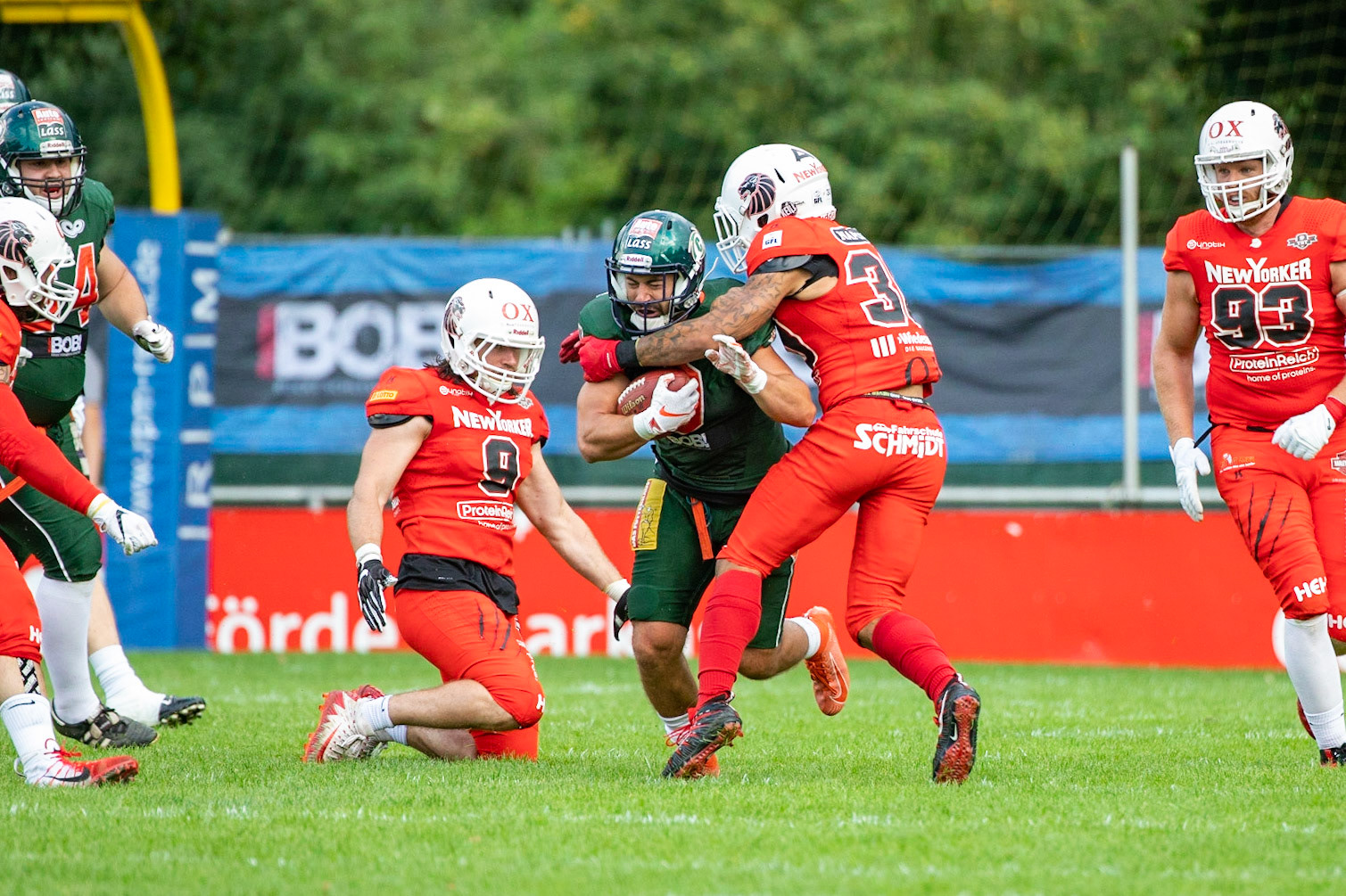 9 Aaron Bruning (grün) gegen 9Roni Salonen, 30 Jamaal White und 93 Julian Völker (von links rot) im Spiel der Kiel Baltic Hurricanes (grün) gegen die Braunschweig New Yorker Lions (rot) am 03.08.19 im Kilia Stadion in Kiel