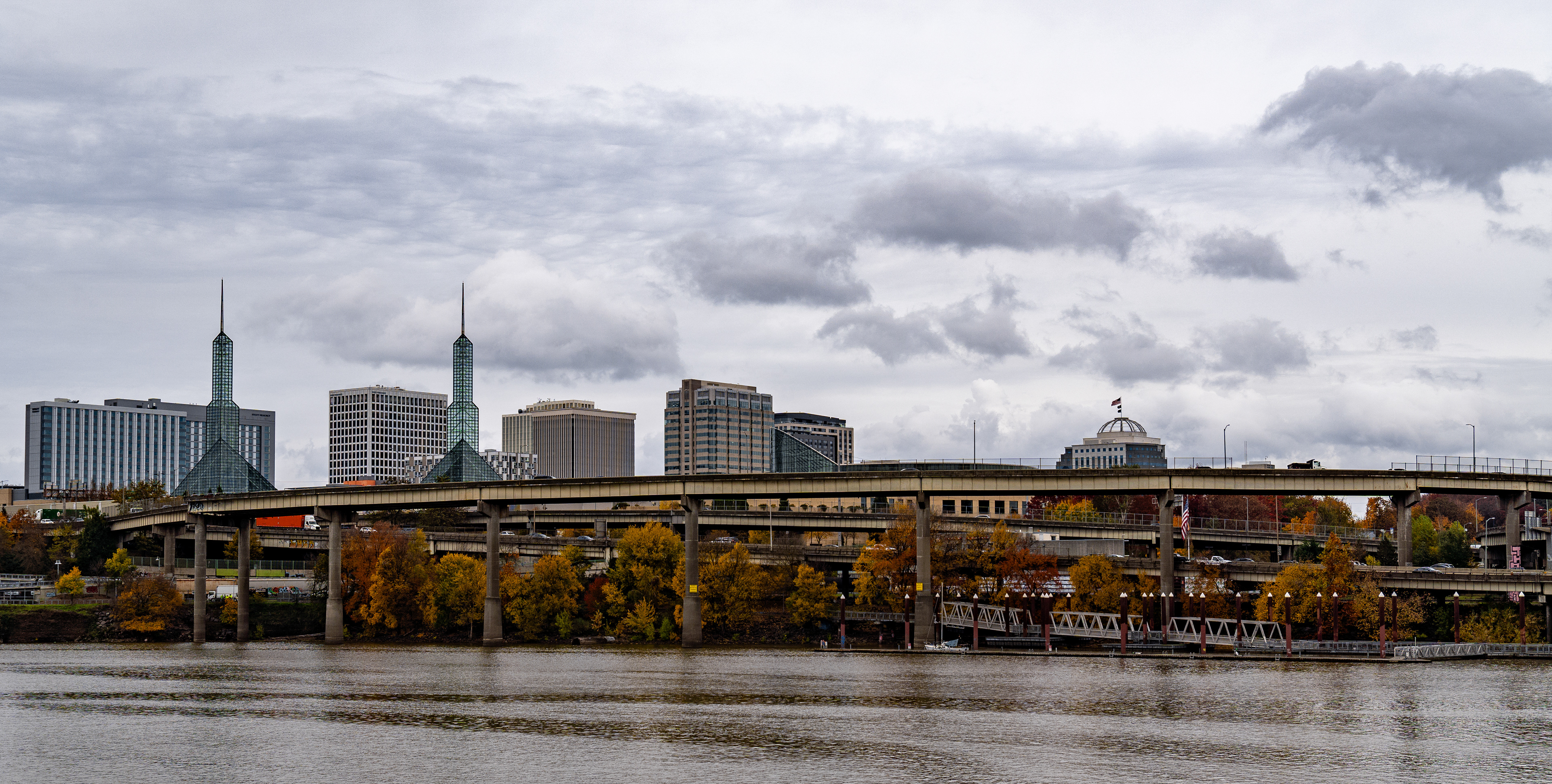 A cloud-covered skyline of Portland looking over the Willamette River. The Oregon Convention Center is seen peeking over a busy I-5.