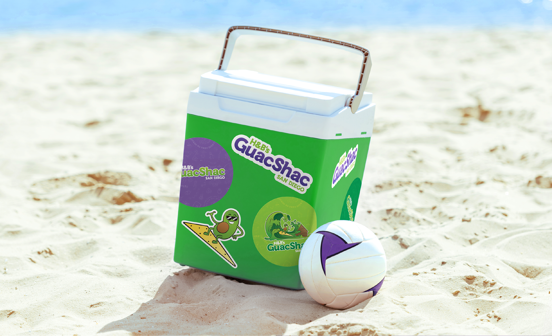 Photo of a cooler with multiple Guac Shac stickers. Cooler and volleyball are on beach sand (designs by Kat Vochoska).