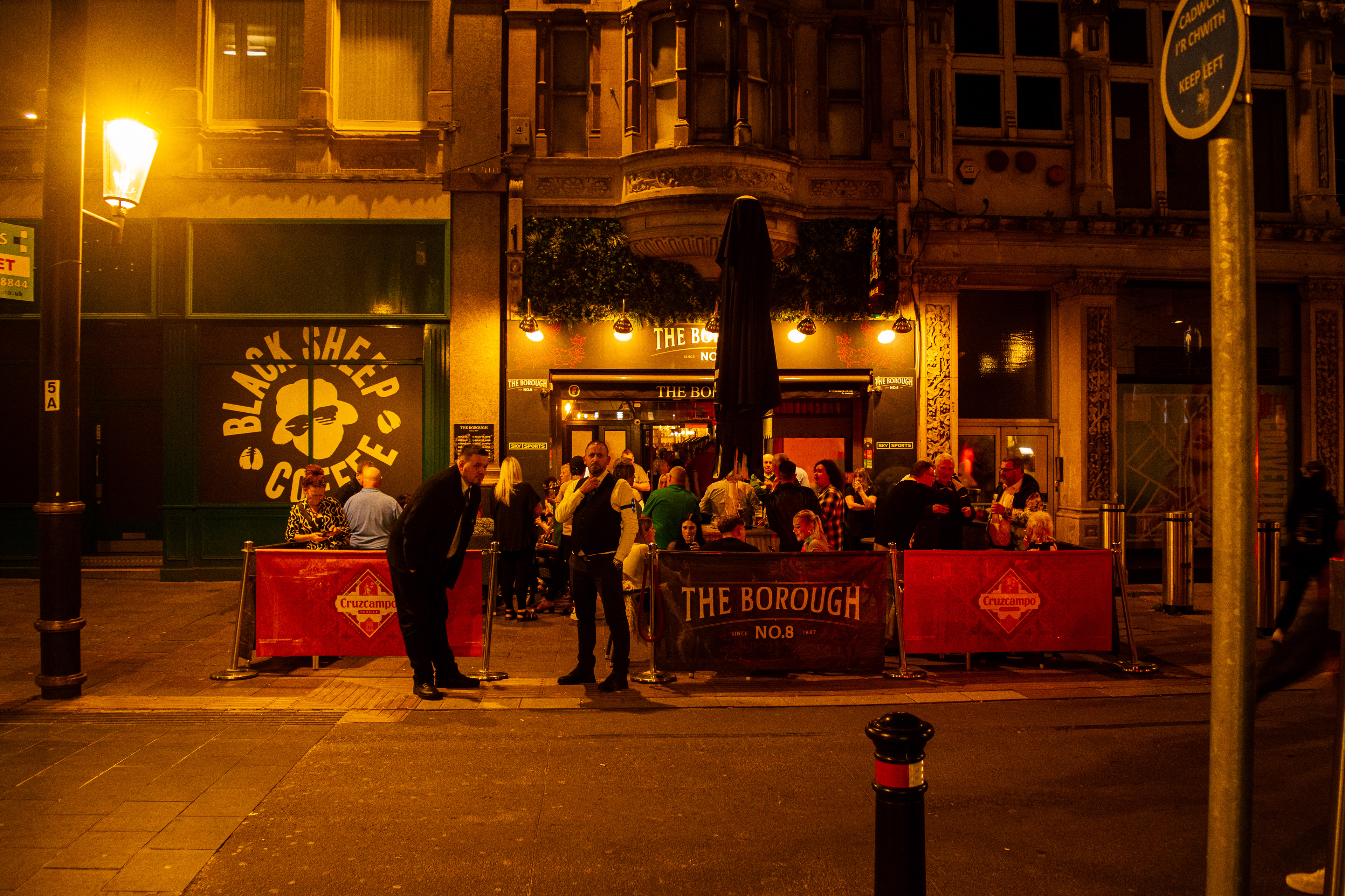 The bouncer looks at the camera while people are having fun in the outdoor section of the pub.