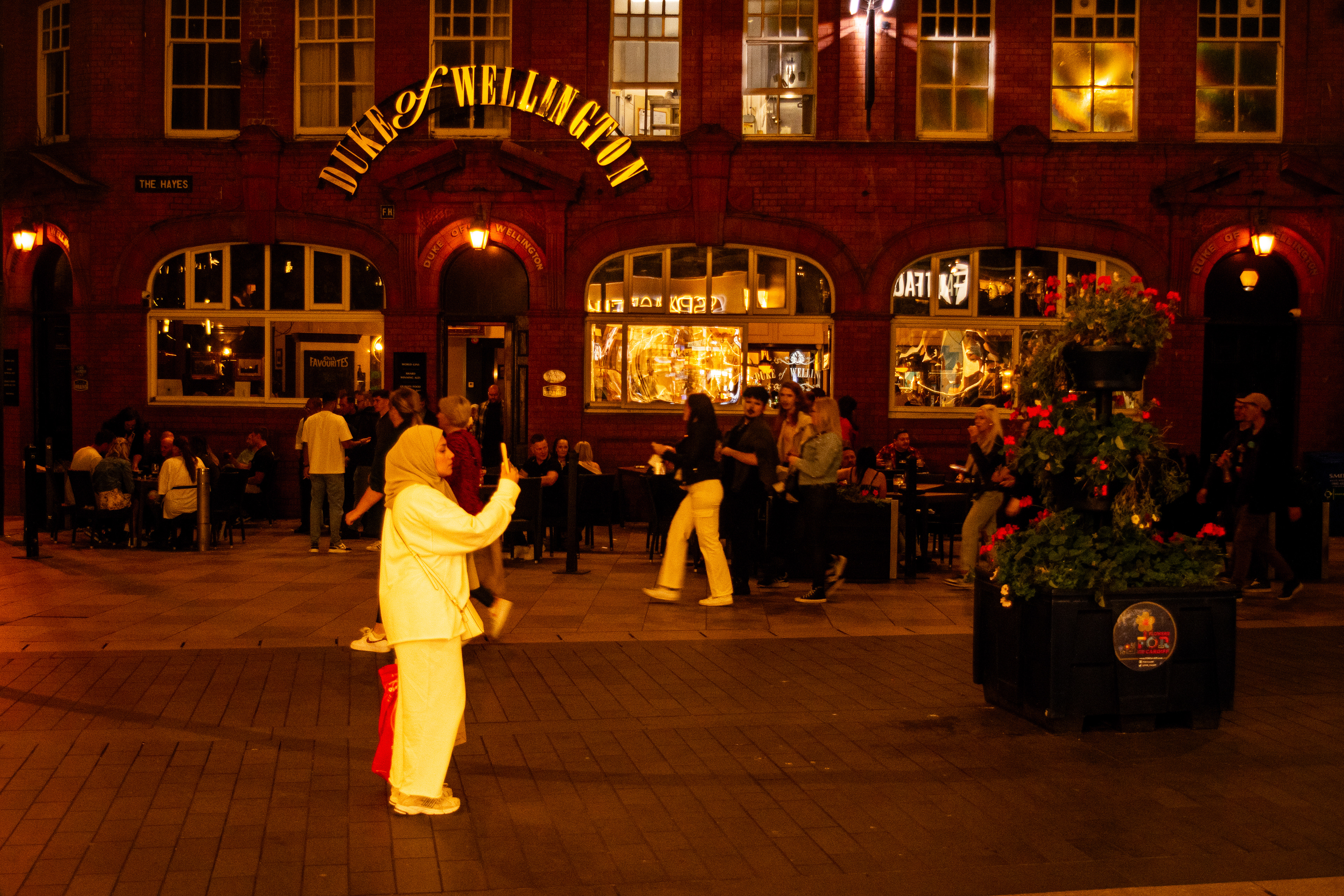 A girl clicks the photo of an illuminated Cardiff City Centre as the people enjoy a weekend night at the pub in the background.