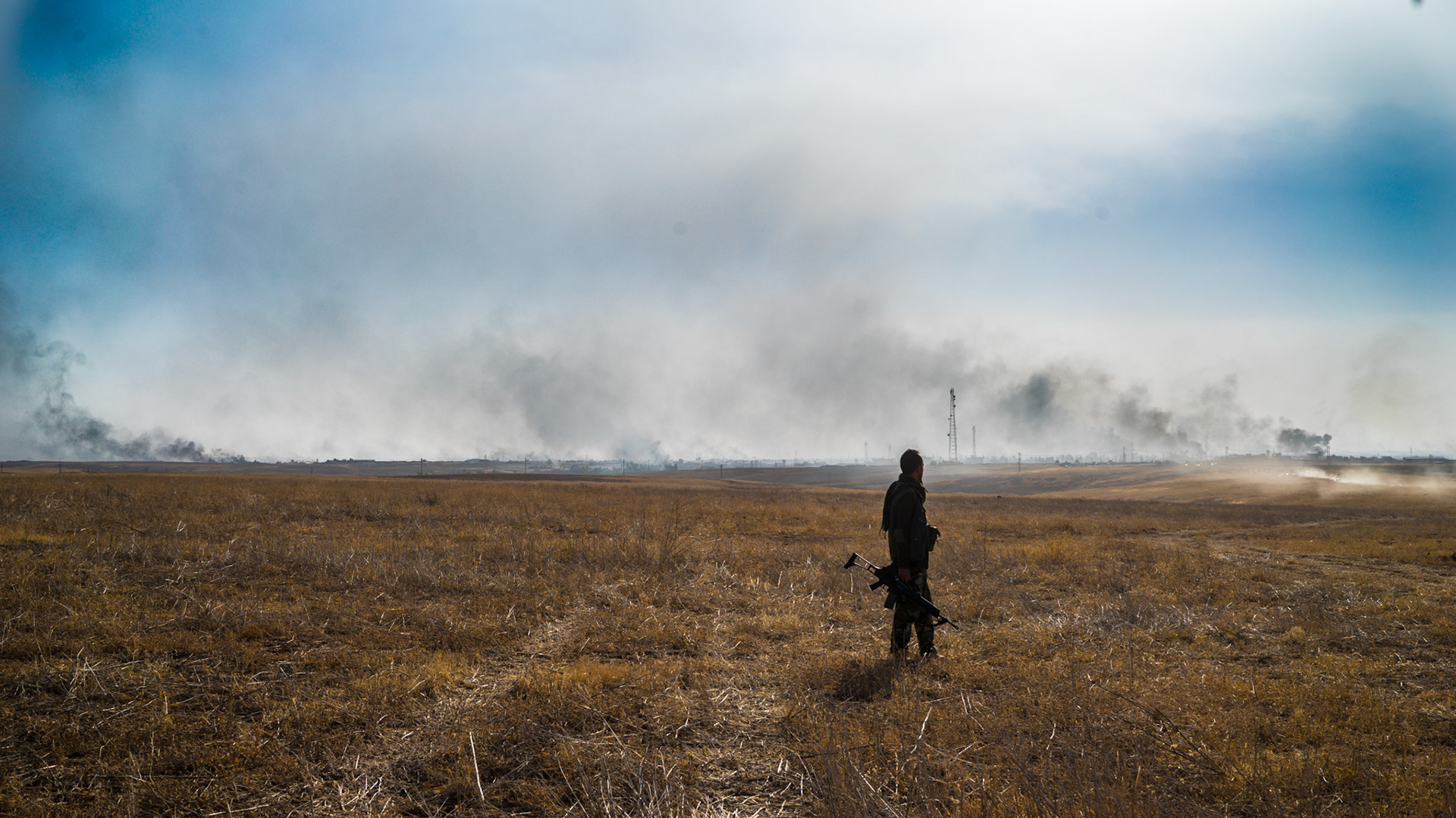 A soldier stands on the battlefield as smoke billows in the distance.