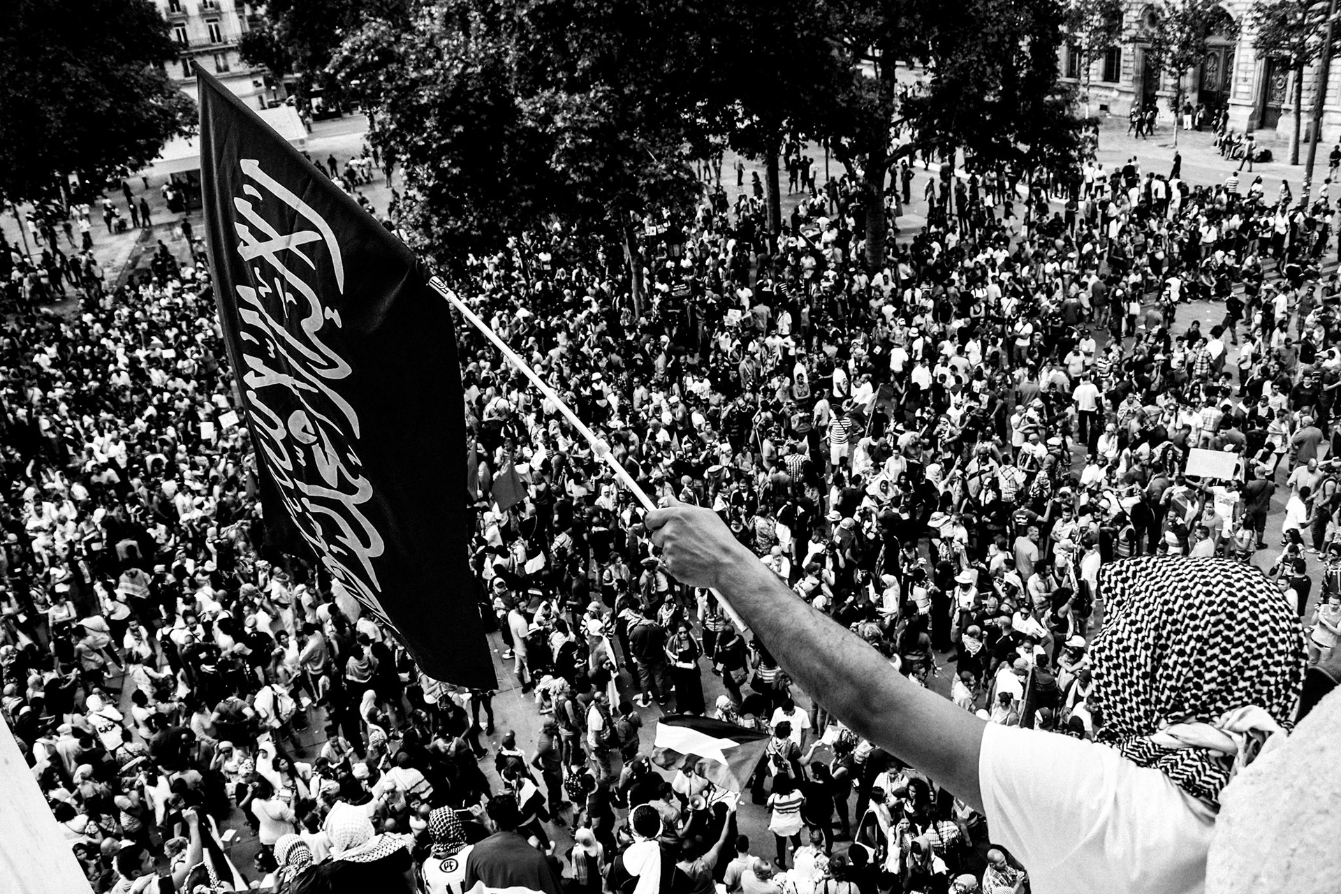 A man stands holding a black flag bearing the Islamic shahada over a crowd of people below