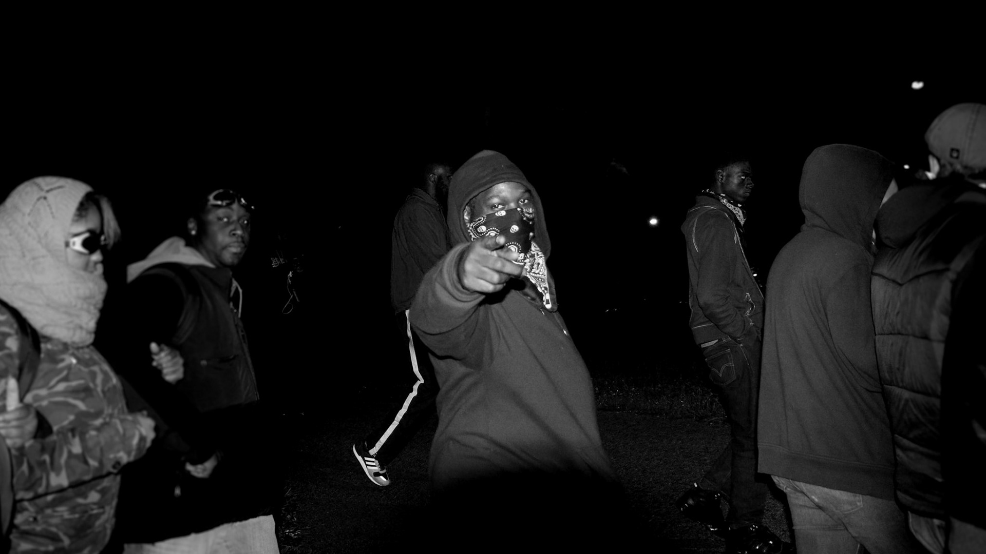A young man wearing a black bandana gestures with his hand in the shape of a gun with his eyes looking directly into the camera