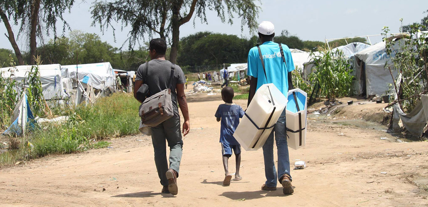 Designer Jason Robinson, UNICEF worker and child walk in IDP camp with Adolescent Kit in Bor, South Sudan.