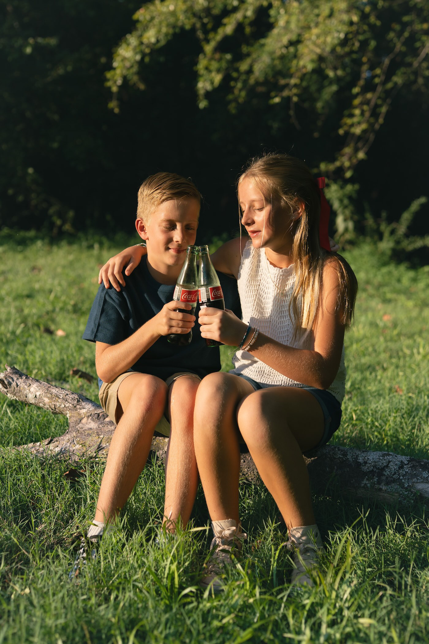Brother and sister drinking Coca-Cola while sitting together on a log outdoors