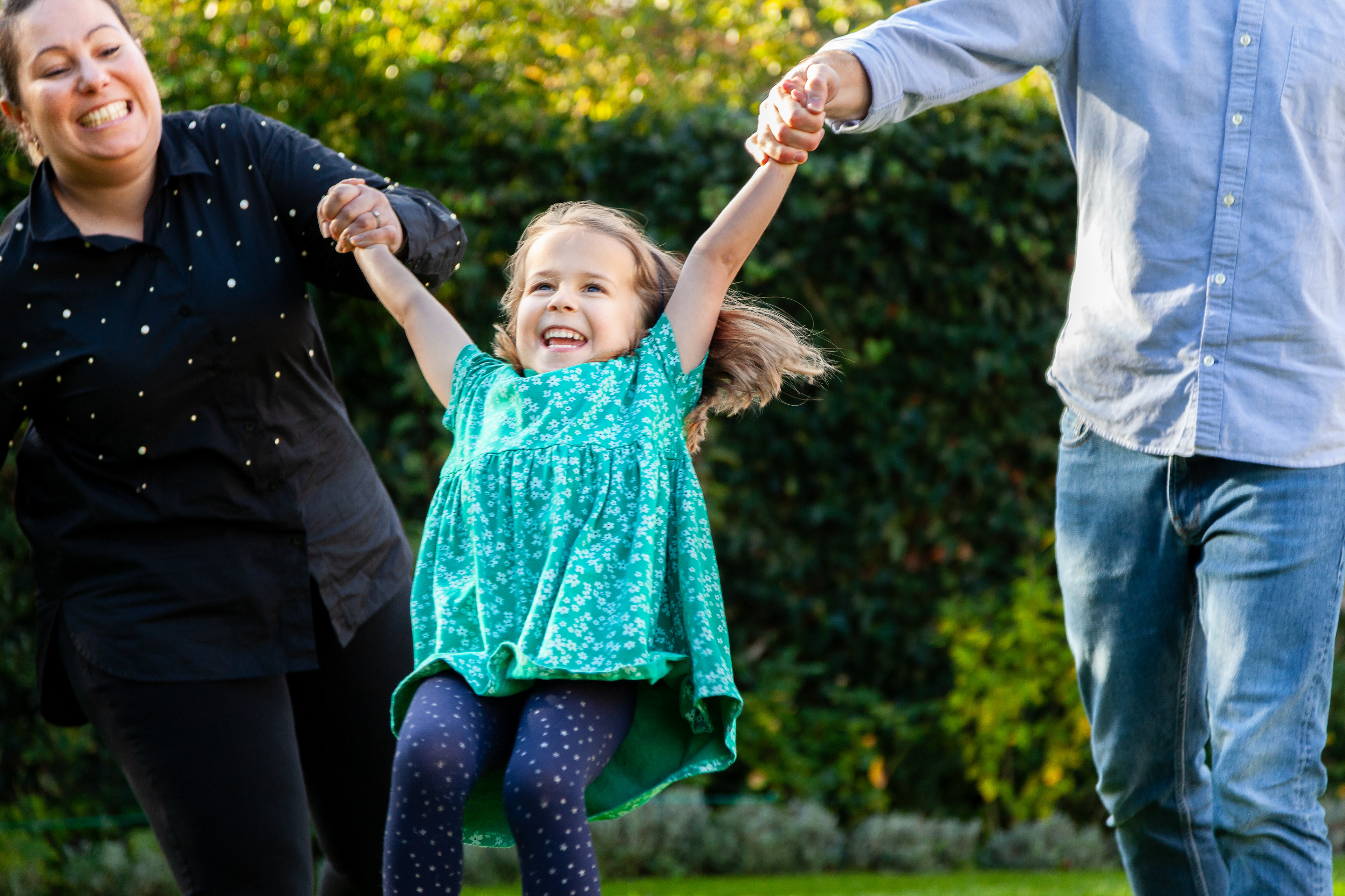 Mom and dad are swinging a little girl in a green dress. Dad's head is not visible. Mom and daughter are laughing. Portrait photographer Hampshire, Surrey, Berkshire. Family, children, headshots, branding.