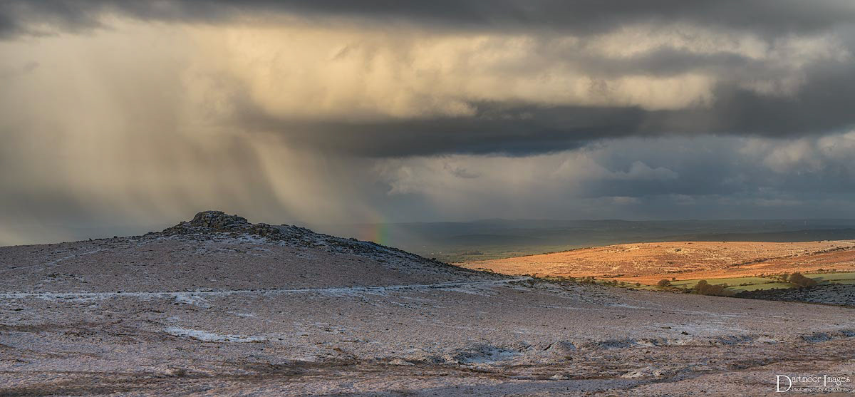 A cold, stormy winters morning on Dartmoor National Park in Devon reveals a light dusting of snow that has fallen overnight in the area of Merrivale, just to the east of Princetown and the infamous Dartmoor prison. Dark and moody storm clouds still dominate the sky bringing with them further snow showers but the occasional break in the clouds enables weak sun to highlight patches of the landscape and in this instance cause a small rainbow to form over the granite peak of Kings Tor