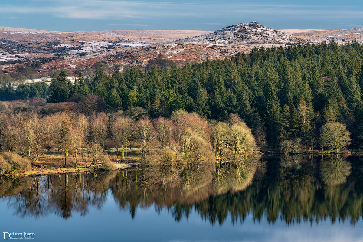 A light dusting of snow lies on and around Down Tor on Dartmoor National Park with the still waters of Burrator Reservoir reflecting the trees and plantations that surround the lake.